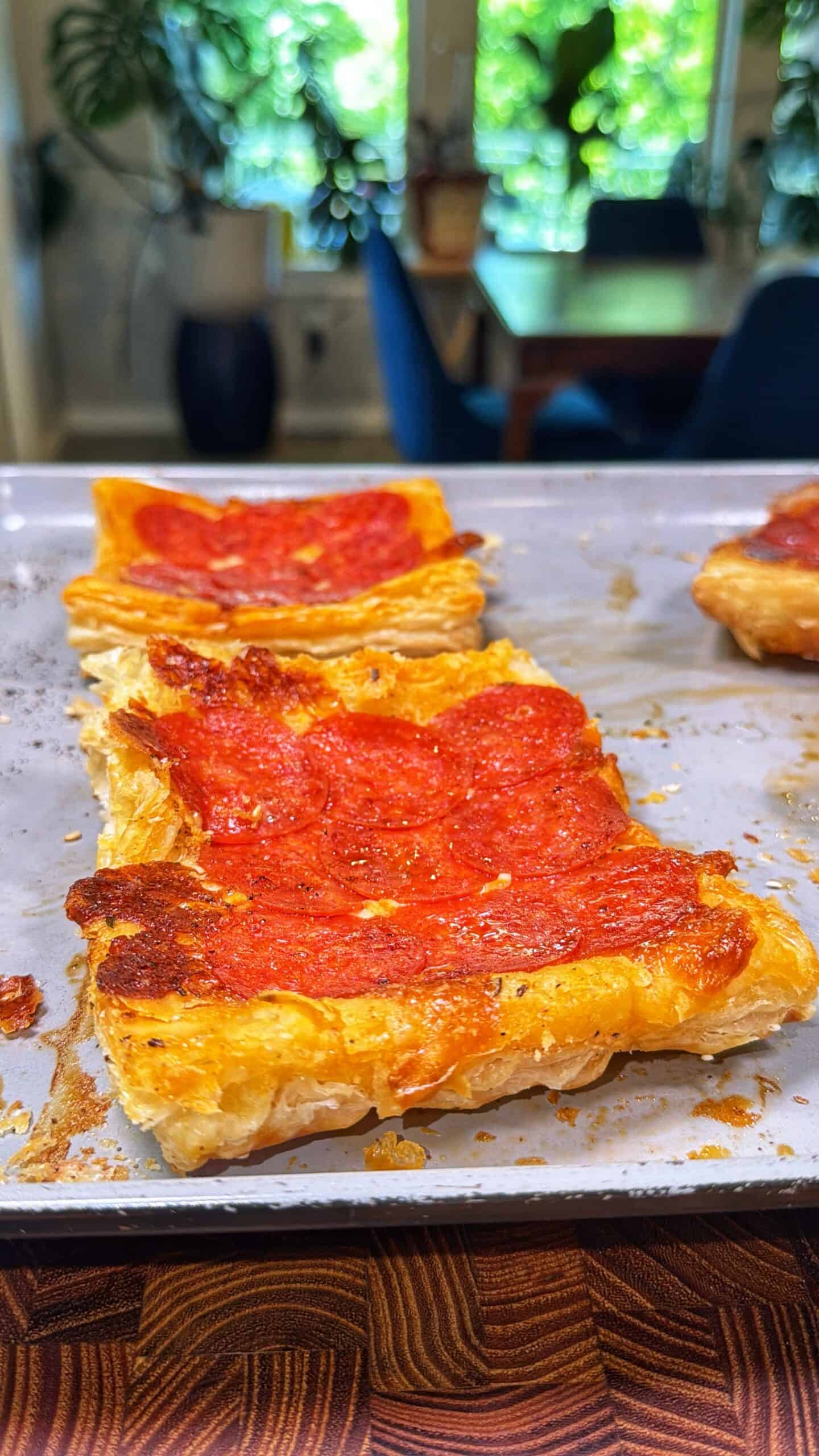 Rectangular puff pastry topped with melted cheese and pepperoni slices on a baking tray. The background shows a kitchen or dining area with greenery and blue chairs visible.
