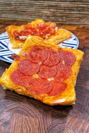 A close-up of a slice of pepperoni pizza with a golden crust on a wooden surface, with another pizza slice topped with tomato sauce in the background on a patterned plate.