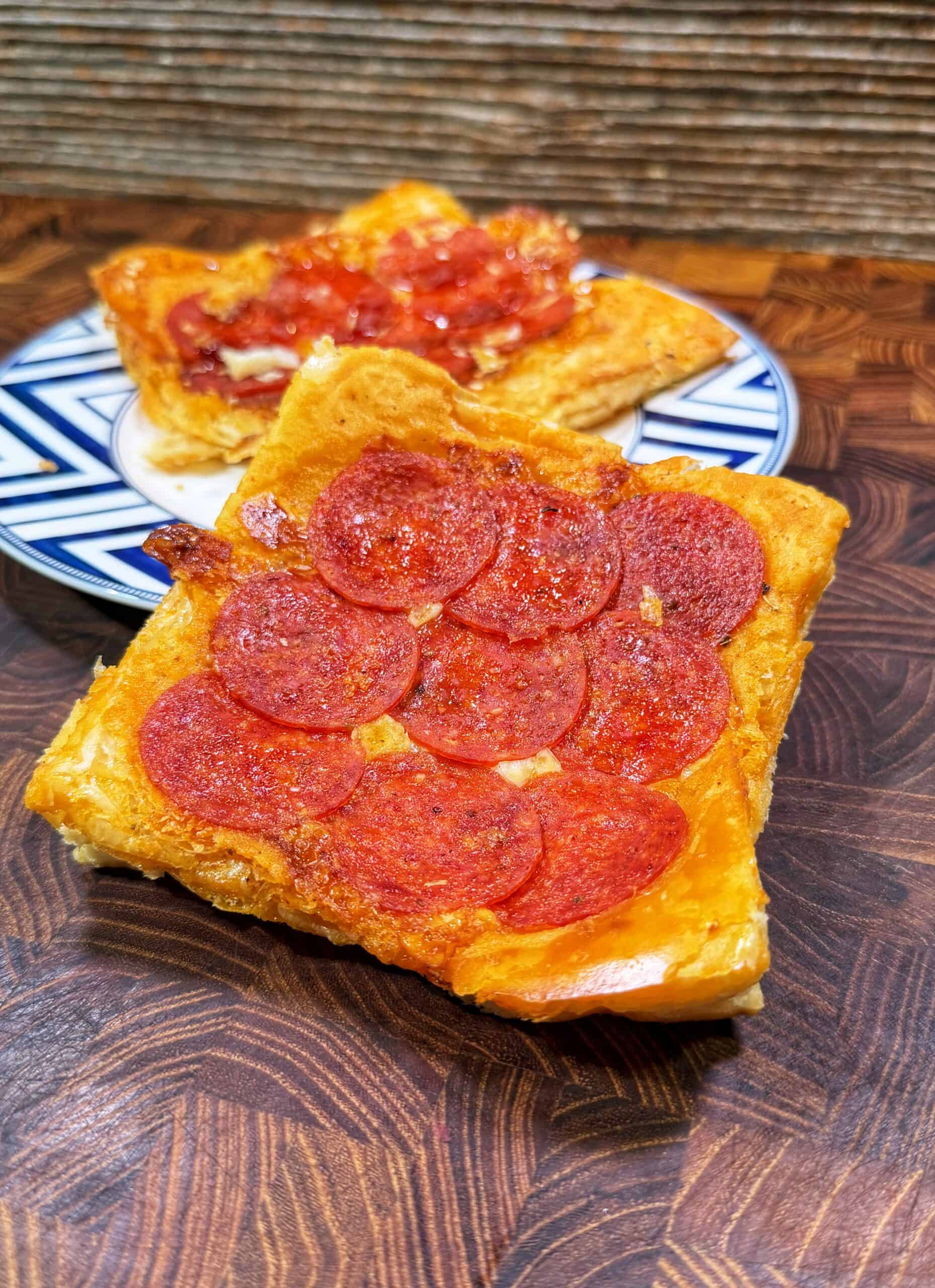 A close-up of a slice of pepperoni pizza with a golden crust on a wooden surface, with another pizza slice topped with tomato sauce in the background on a patterned plate.