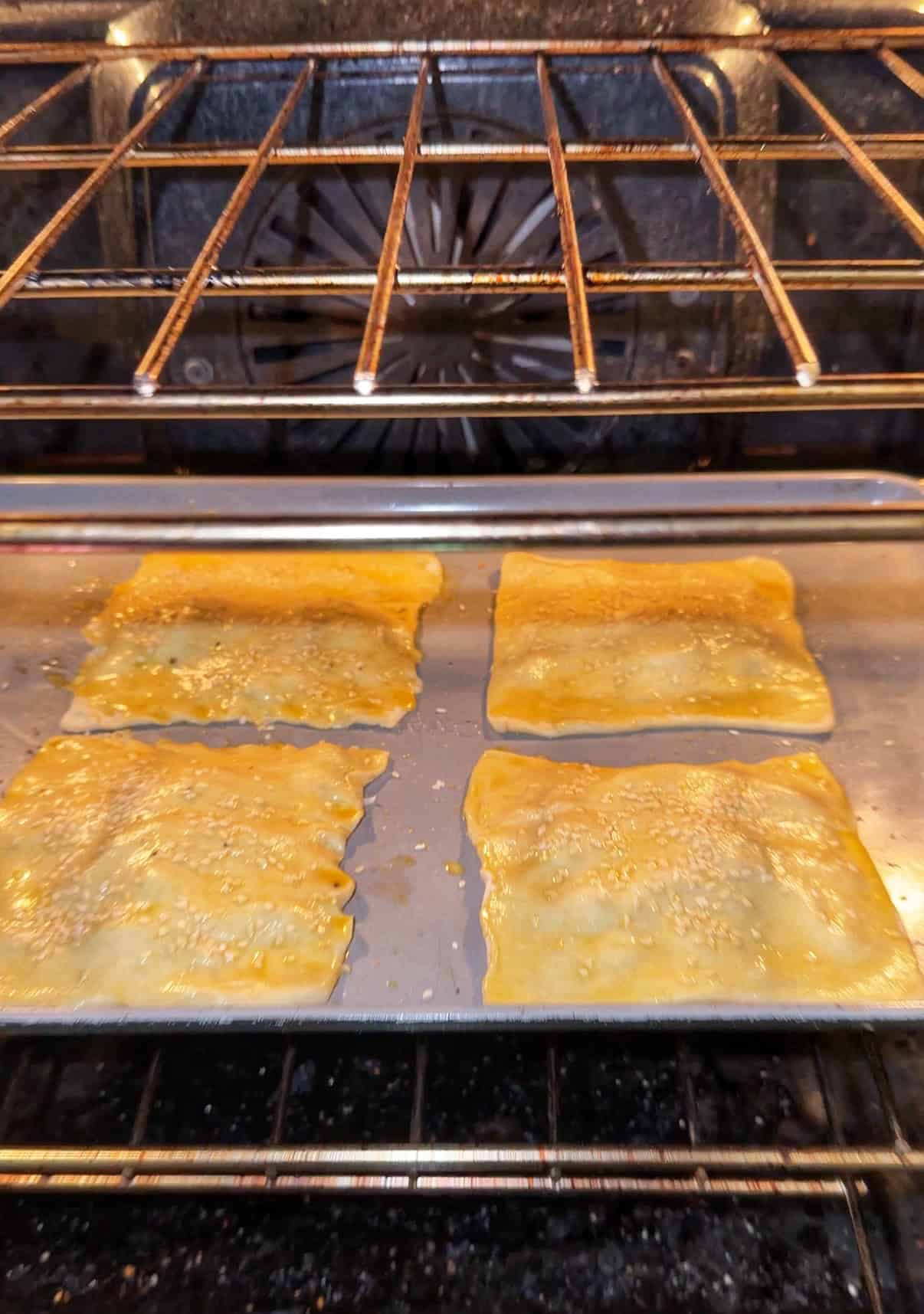 Four square pastries bake on a metal tray inside an oven. The pastries have a golden, shiny surface and are sprinkled with what appears to be sesame seeds. The oven racks and back fan are visible.