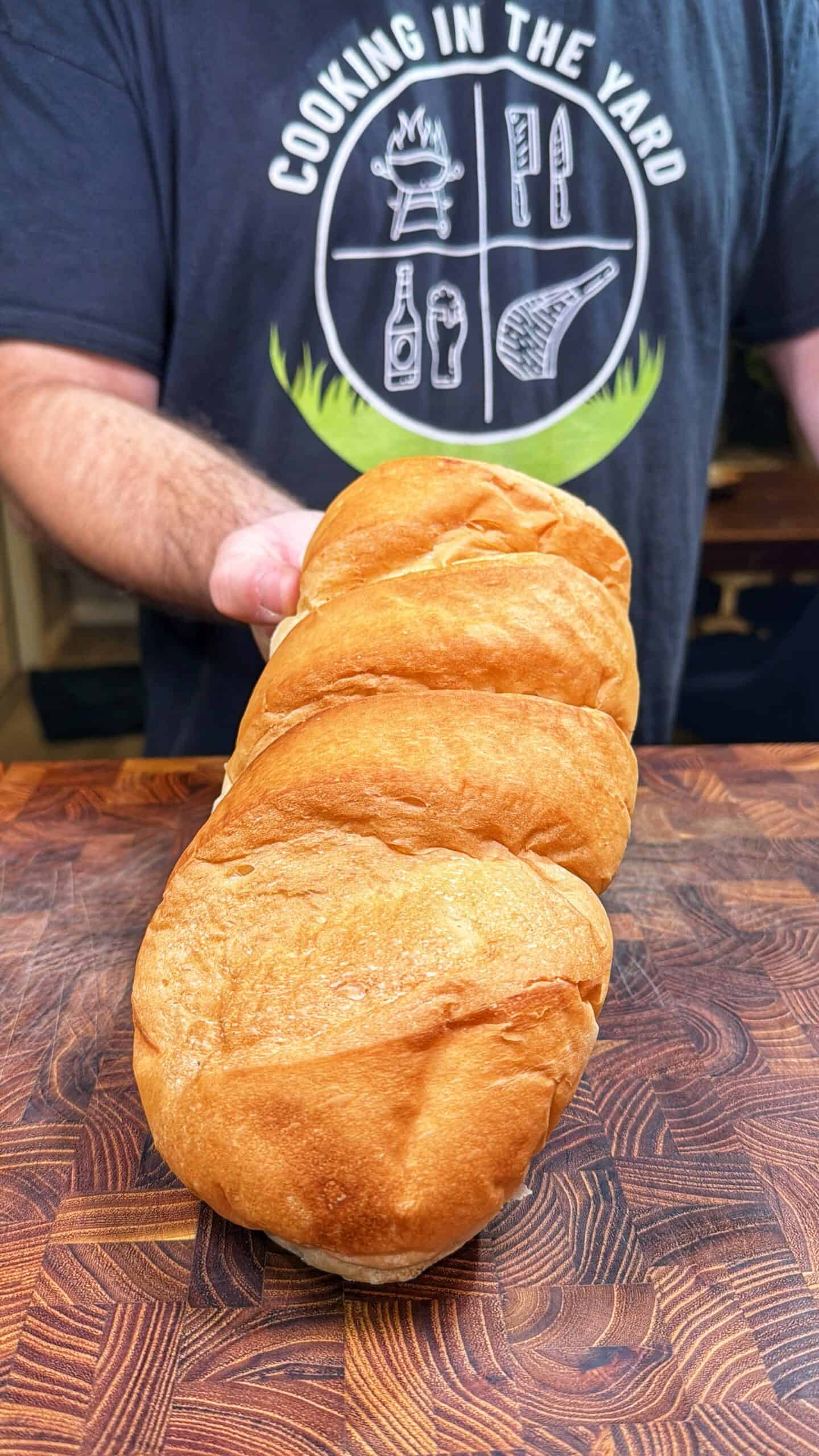 A person wearing a Cooking In The Yard shirt holds a loaf of braided bread on a wooden cutting board.