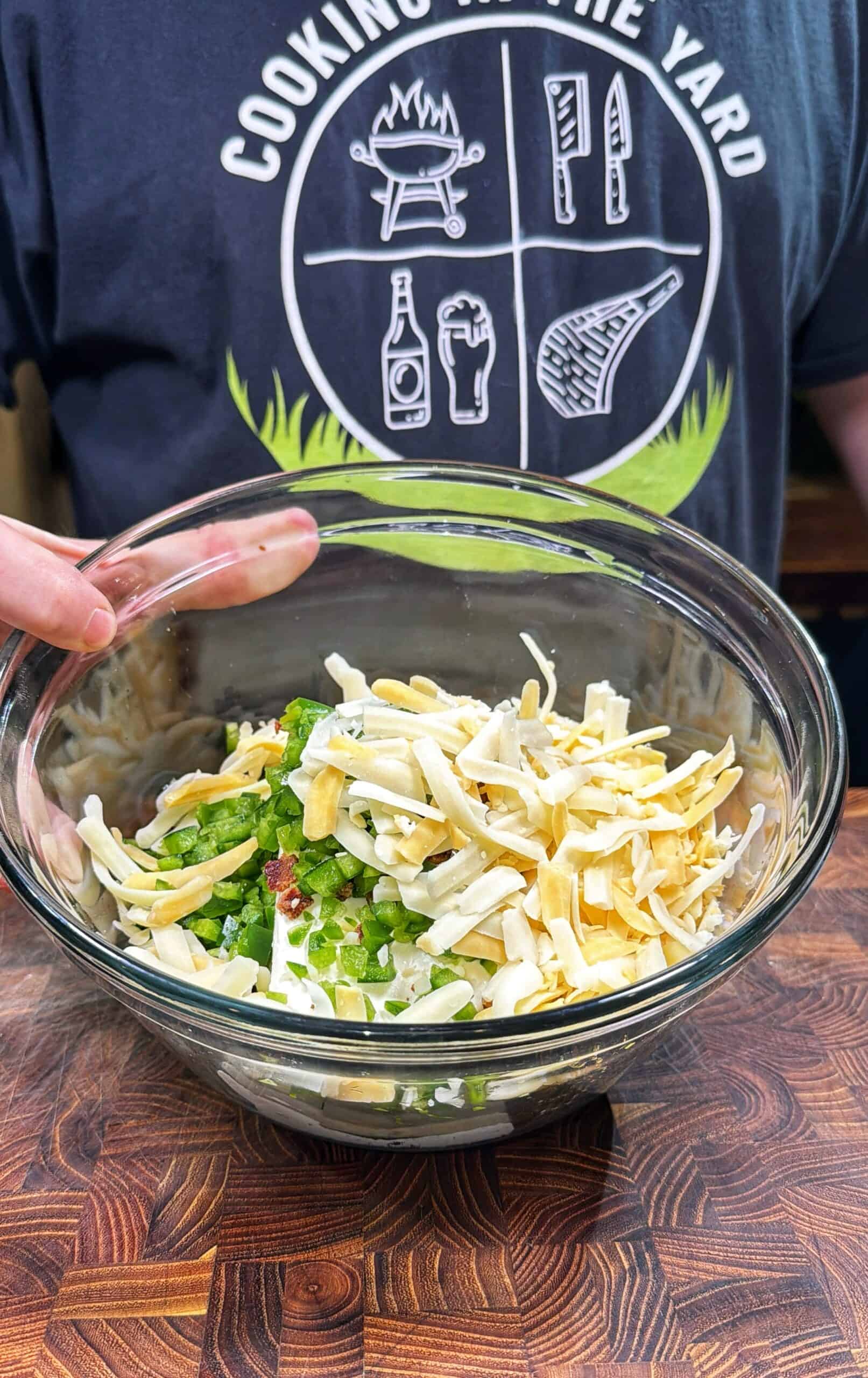 A person holding a glass bowl filled with shredded cheese, chopped green onions, and bacon bits on a wooden surface. The person is wearing a dark shirt with grilling icons and text that reads Cooking in the Yard.