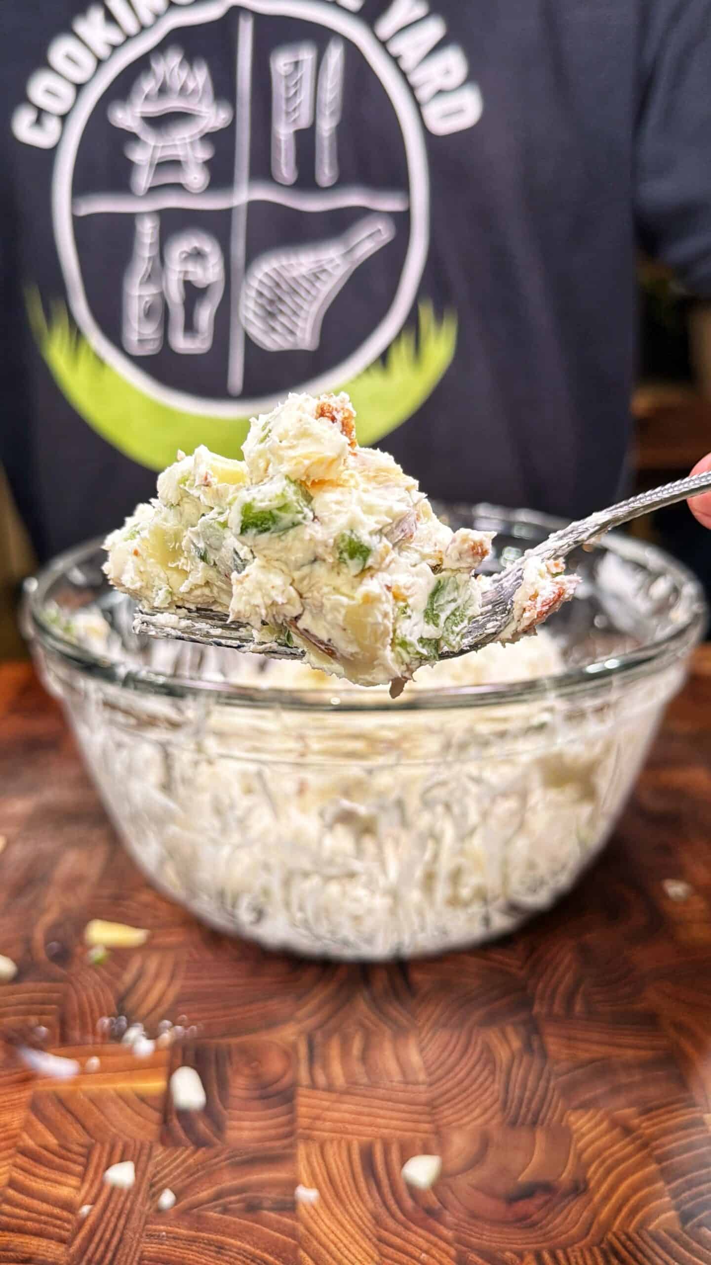 A person holds a spoonful of creamy potato salad with herbs above a glass bowl of the same salad. The person is wearing a dark shirt with a cooking-themed graphic.