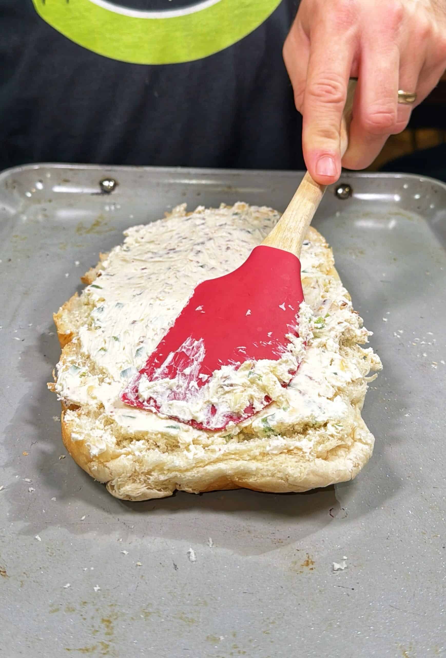 A person spreads a creamy mixture onto a halved loaf of bread using a red spatula on a baking tray.