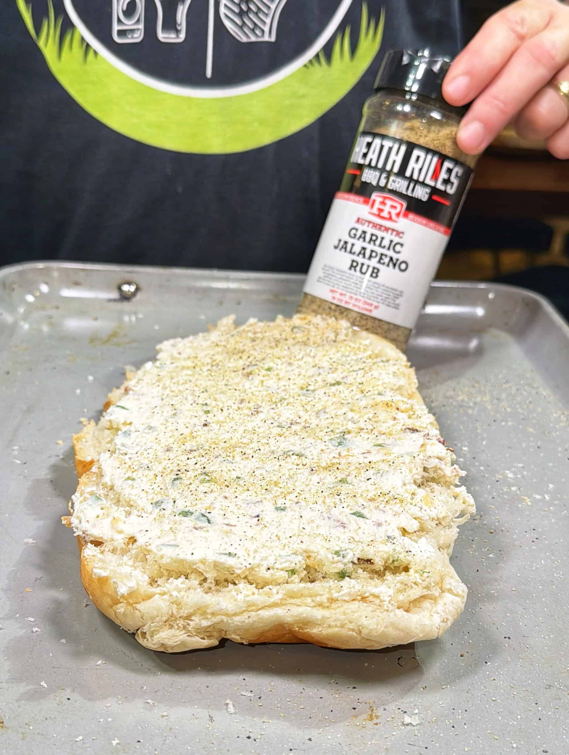 A hand sprinkles garlic jalapeño rub onto a loaf of bread topped with a creamy spread, all placed on a baking tray. The person wears a dark shirt with kitchen utensils printed on it.