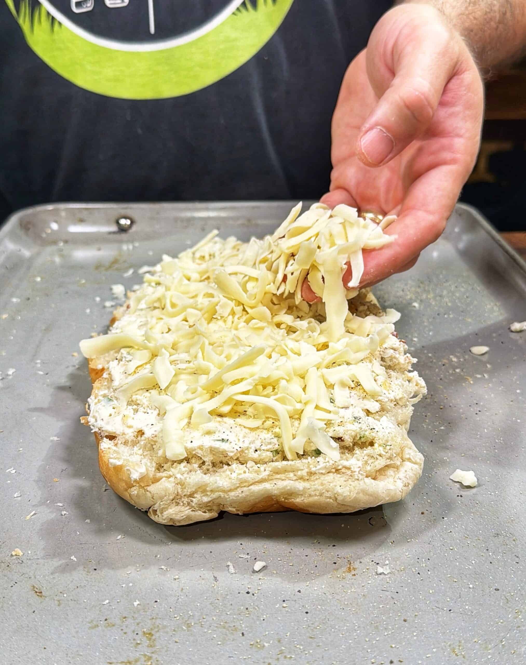 A person sprinkles shredded cheese onto a piece of bread covered with a creamy spread, preparing it on a baking sheet.