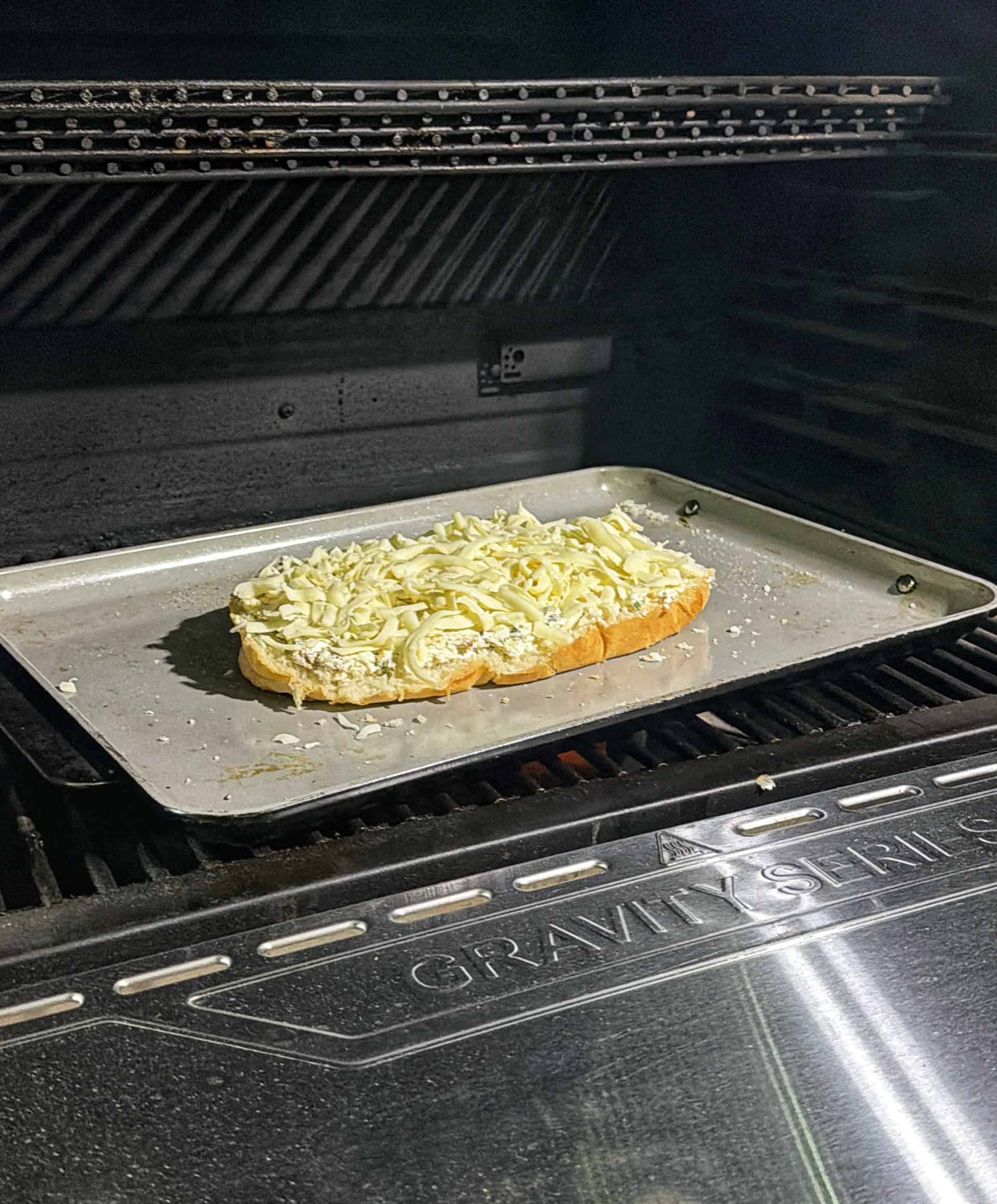 A slice of bread topped with shredded cheese on a metal baking sheet is placed inside a grill or oven for toasting.