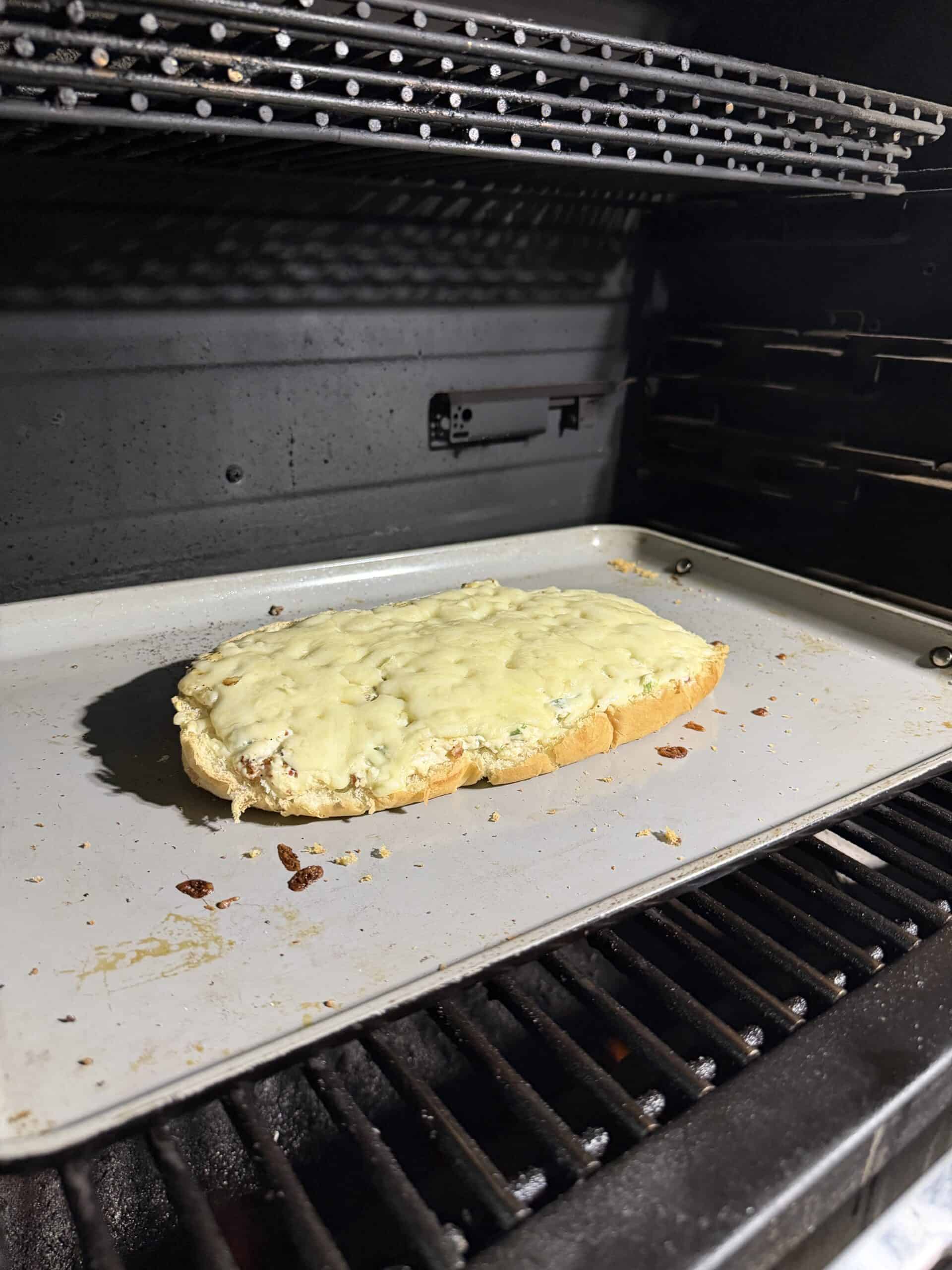 A slice of bread topped with melted cheese sits on a baking sheet inside an oven, with the cheese slightly browned and the bread edges toasted.