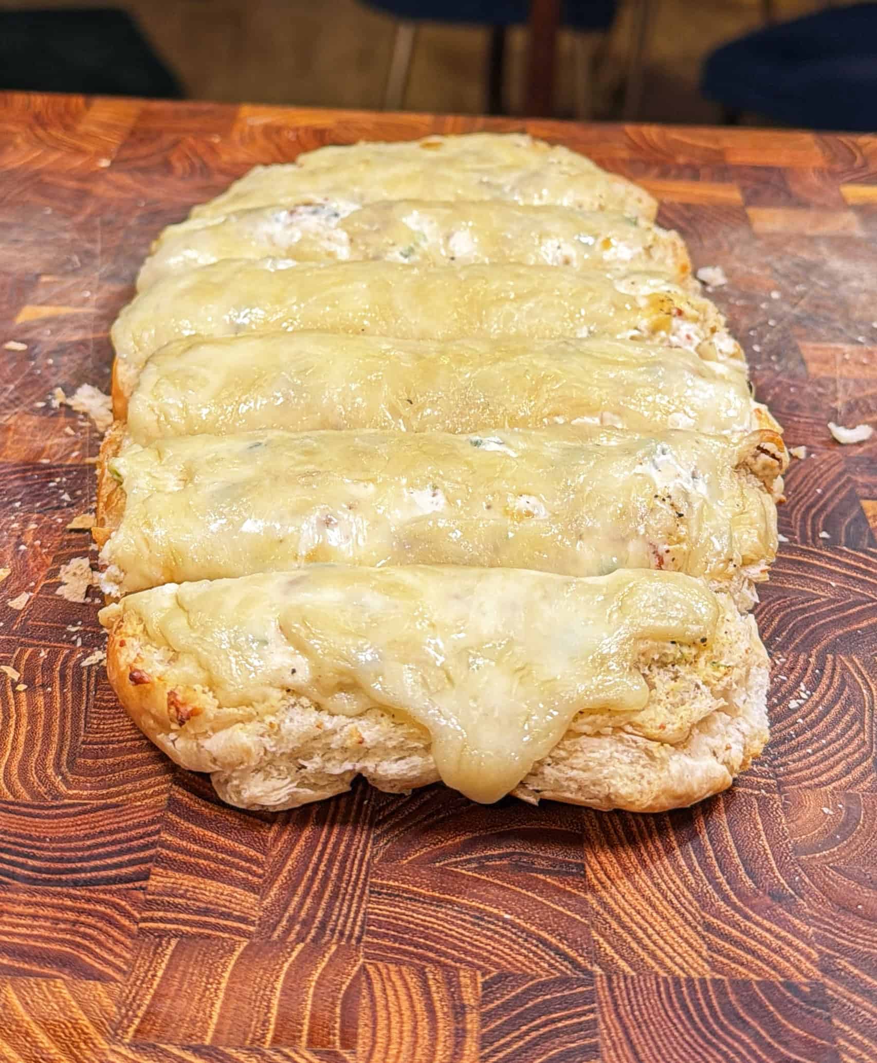 A loaf of bread sliced into sections and topped with melted cheese, sitting on a patterned wooden cutting board.