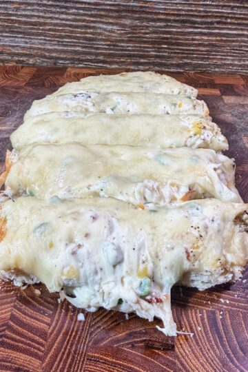 A sliced cheesy bread loaf topped with melted cheese and bits of vegetables, placed on a wooden cutting board with a rustic background.