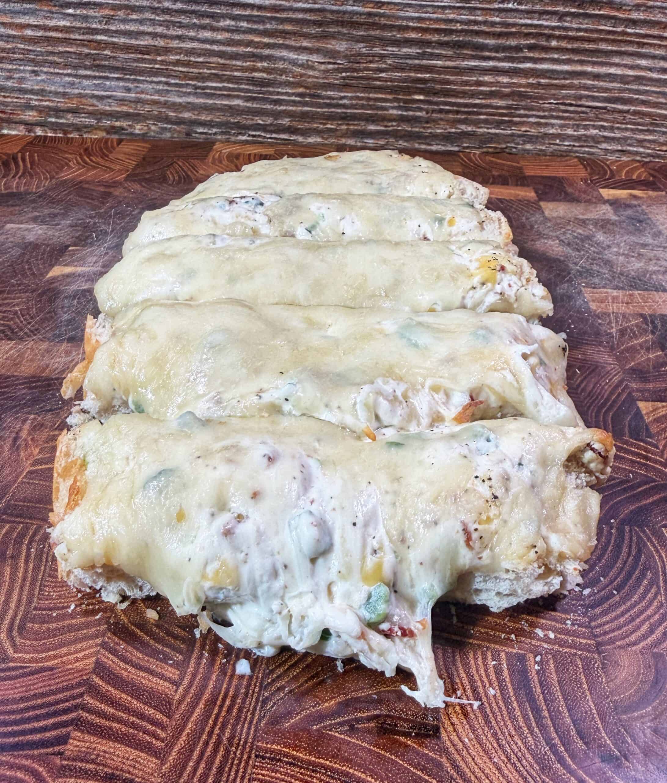 A sliced cheesy bread loaf topped with melted cheese and bits of vegetables, placed on a wooden cutting board with a rustic background.