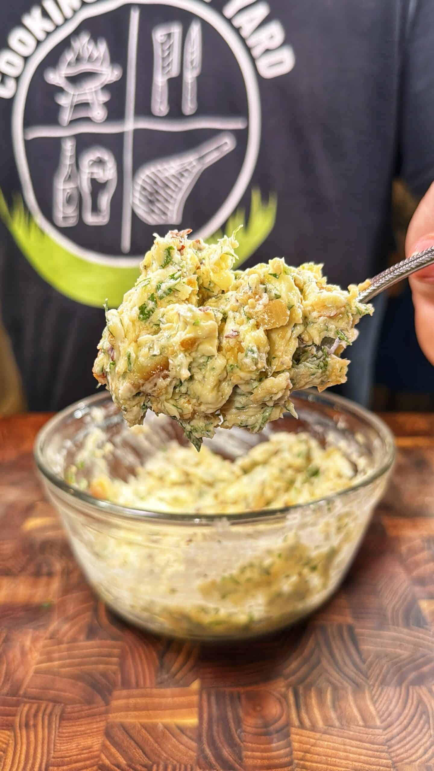 A person holds a spoonful of a chunky, herbed mixture above a glass bowl filled with the same mixture, with a wooden surface and a shirt featuring kitchen utensil graphics in the background.