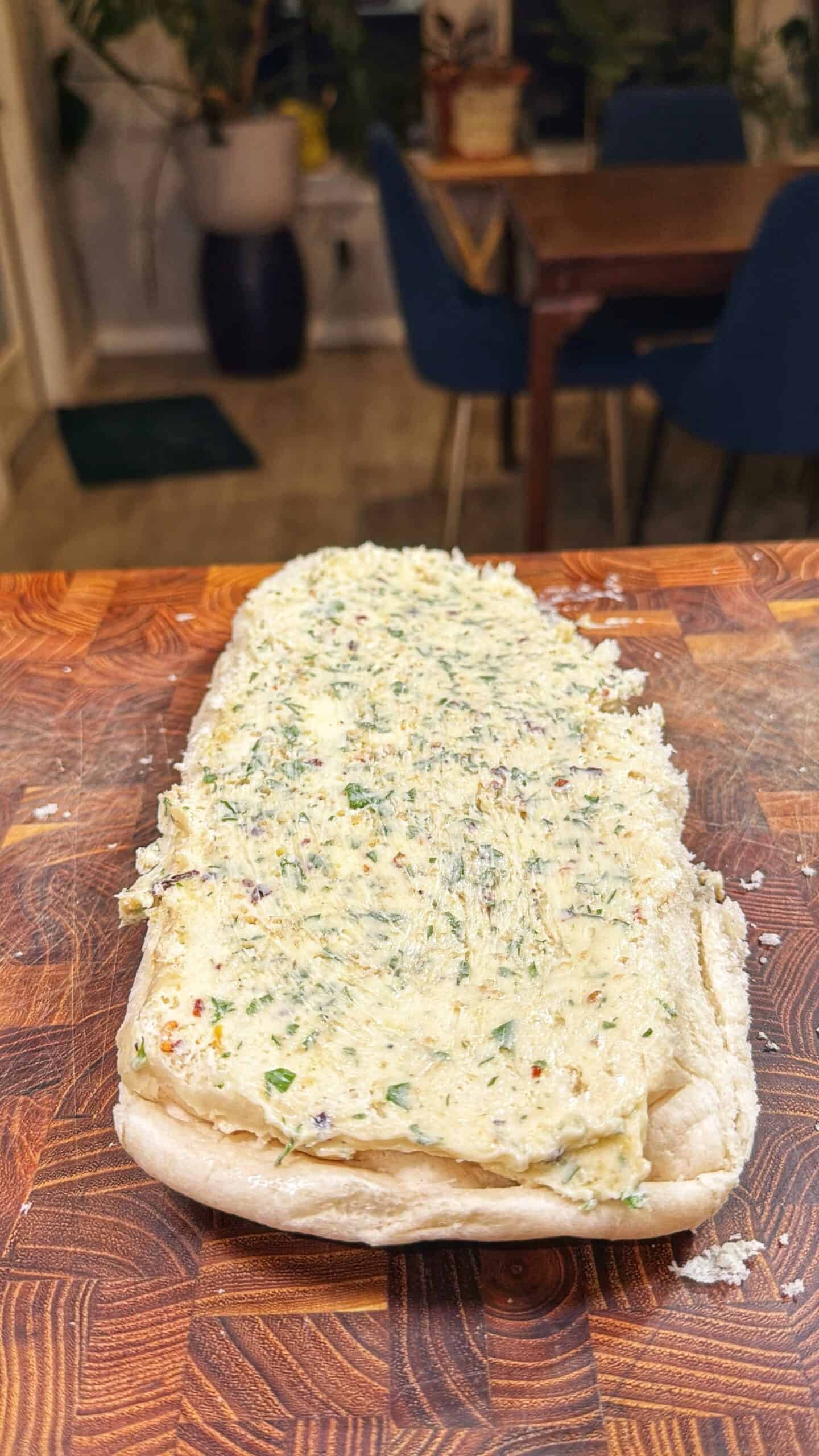 A loaf of bread cut open with a thick layer of herb and spice butter spread on top, resting on a wooden cutting board. A dining table and chairs are visible in the background.