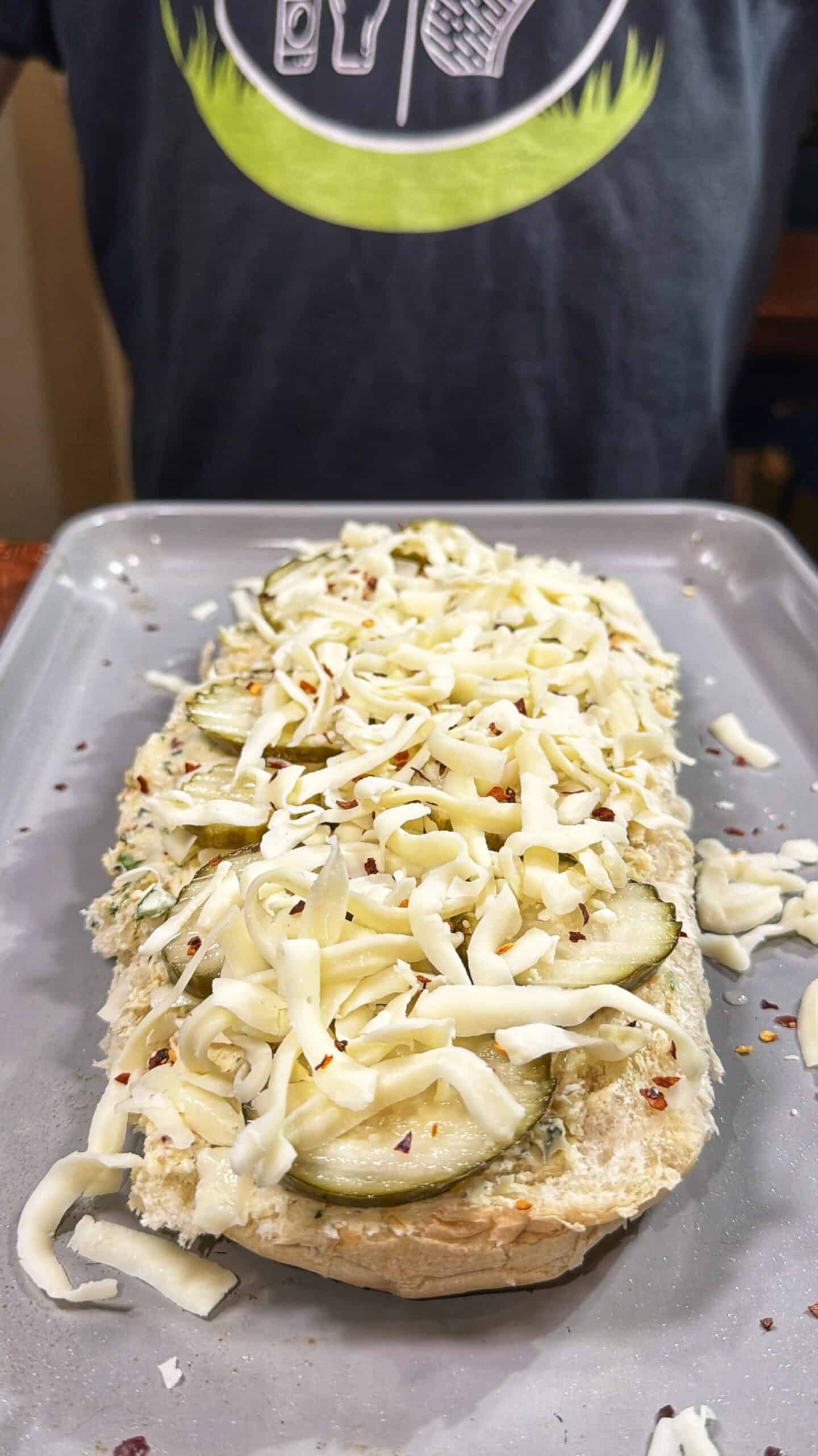 A baking tray holds an open-faced sandwich topped with sliced cucumbers, shredded cheese, and red pepper flakes. In the background, a person wearing a dark shirt with a green design is visible.