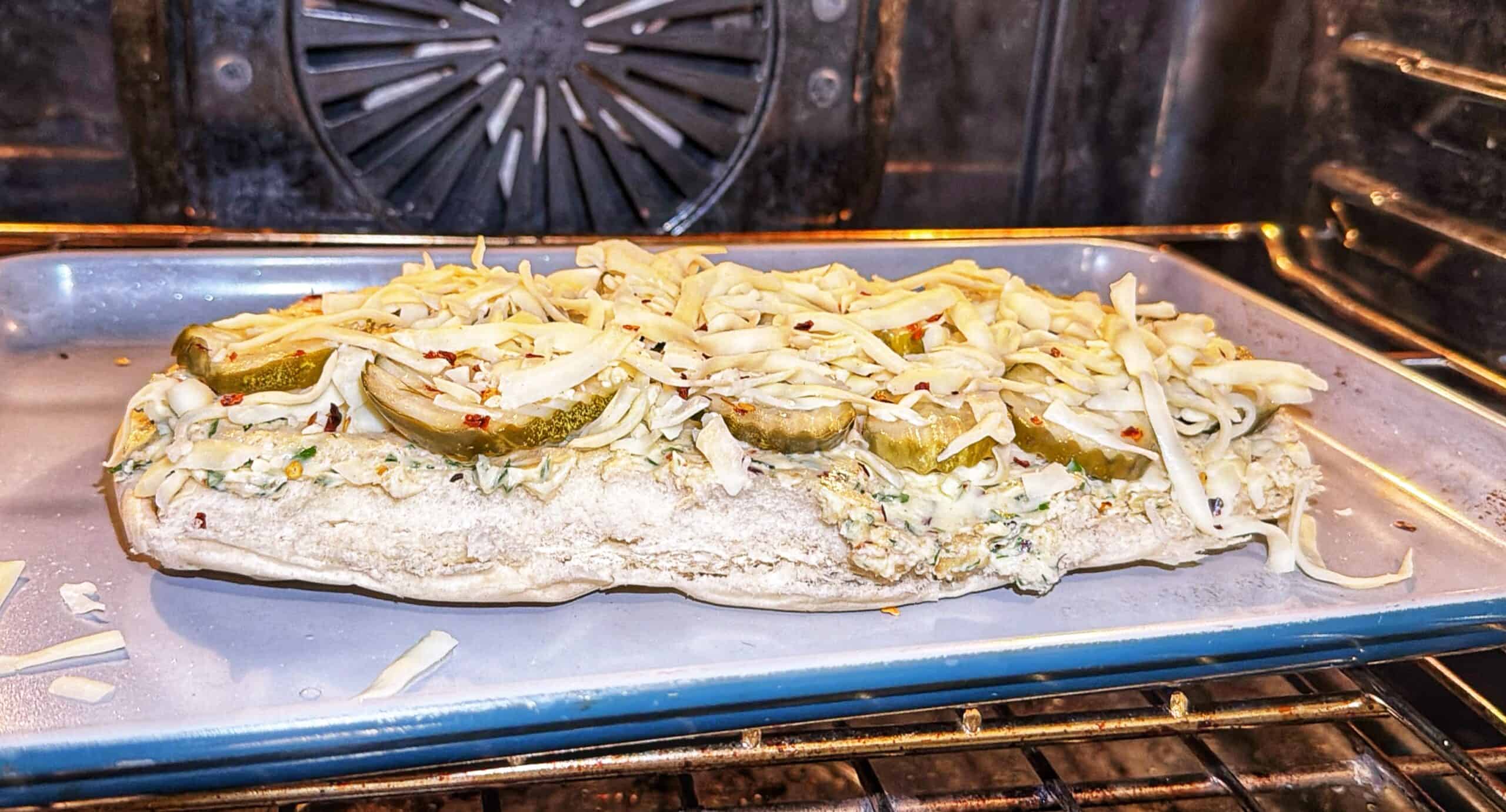 A slice of bread topped with pickles, shredded cheese, and herbs sits on a baking sheet in an oven, melting and toasting under the heat.