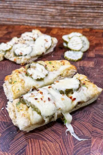 Two pieces of cheesy bread topped with sliced pickles and herbs sit on a wooden board, with extra pickle slices in the background. The melted cheese is slightly browned and garnished with red pepper flakes.
