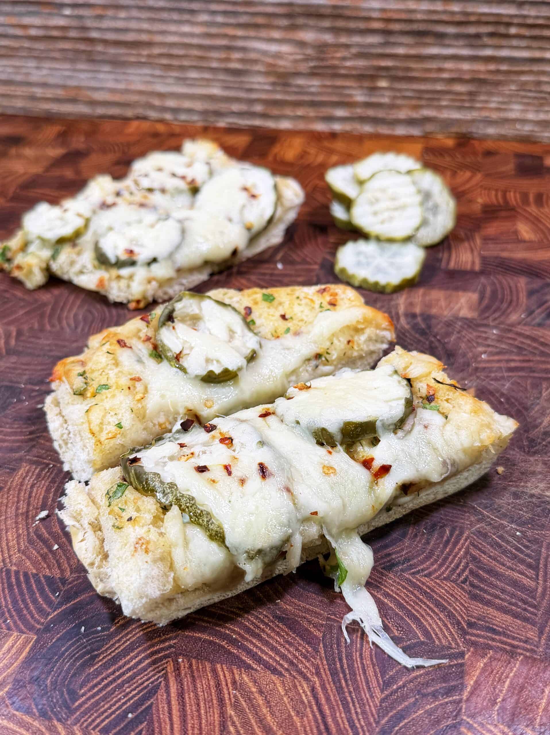 Two pieces of cheesy bread topped with sliced pickles and herbs sit on a wooden board, with extra pickle slices in the background. The melted cheese is slightly browned and garnished with red pepper flakes.