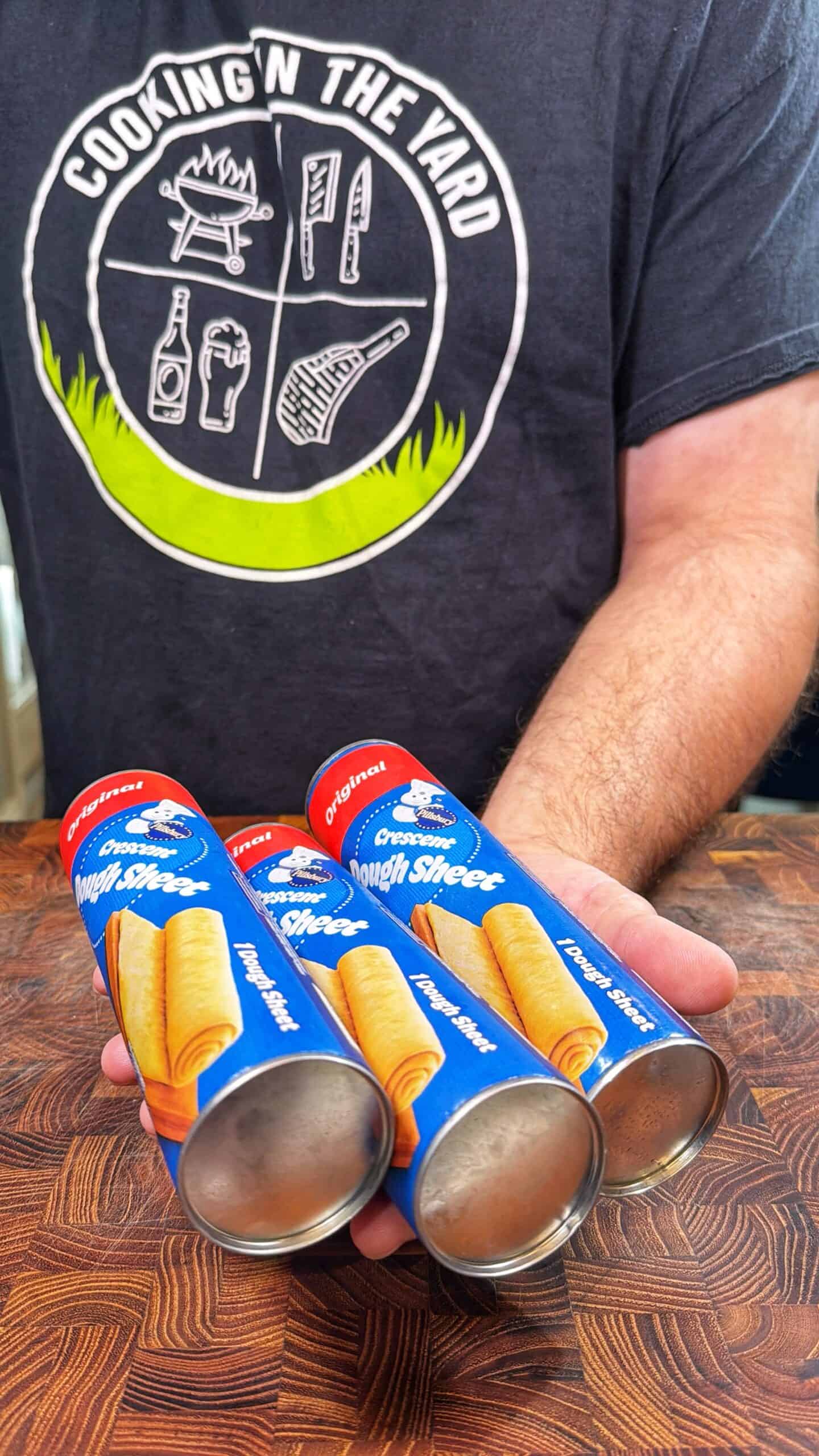 A person wearing a Cooking in the Yard shirt is holding three cans of Great Value Original Dough Sheet on a wooden surface. The cans are blue with images of rolled dough.