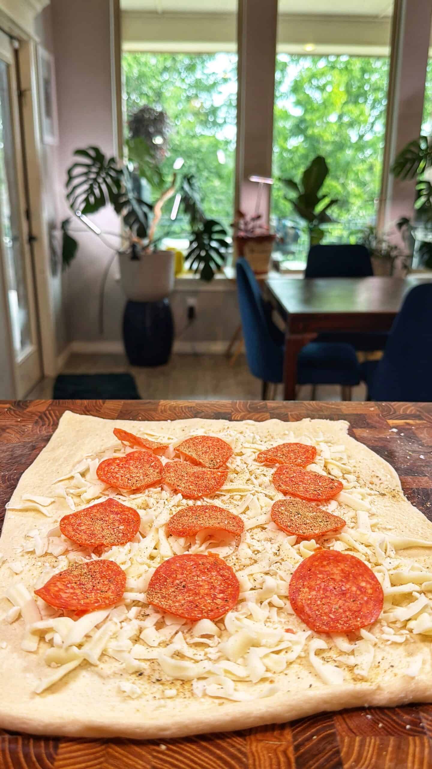 A rectangular sheet of pizza dough topped with shredded cheese and pepperoni sits on a wooden surface. In the background, a dining table, blue chairs, and large green plants are visible by a window.