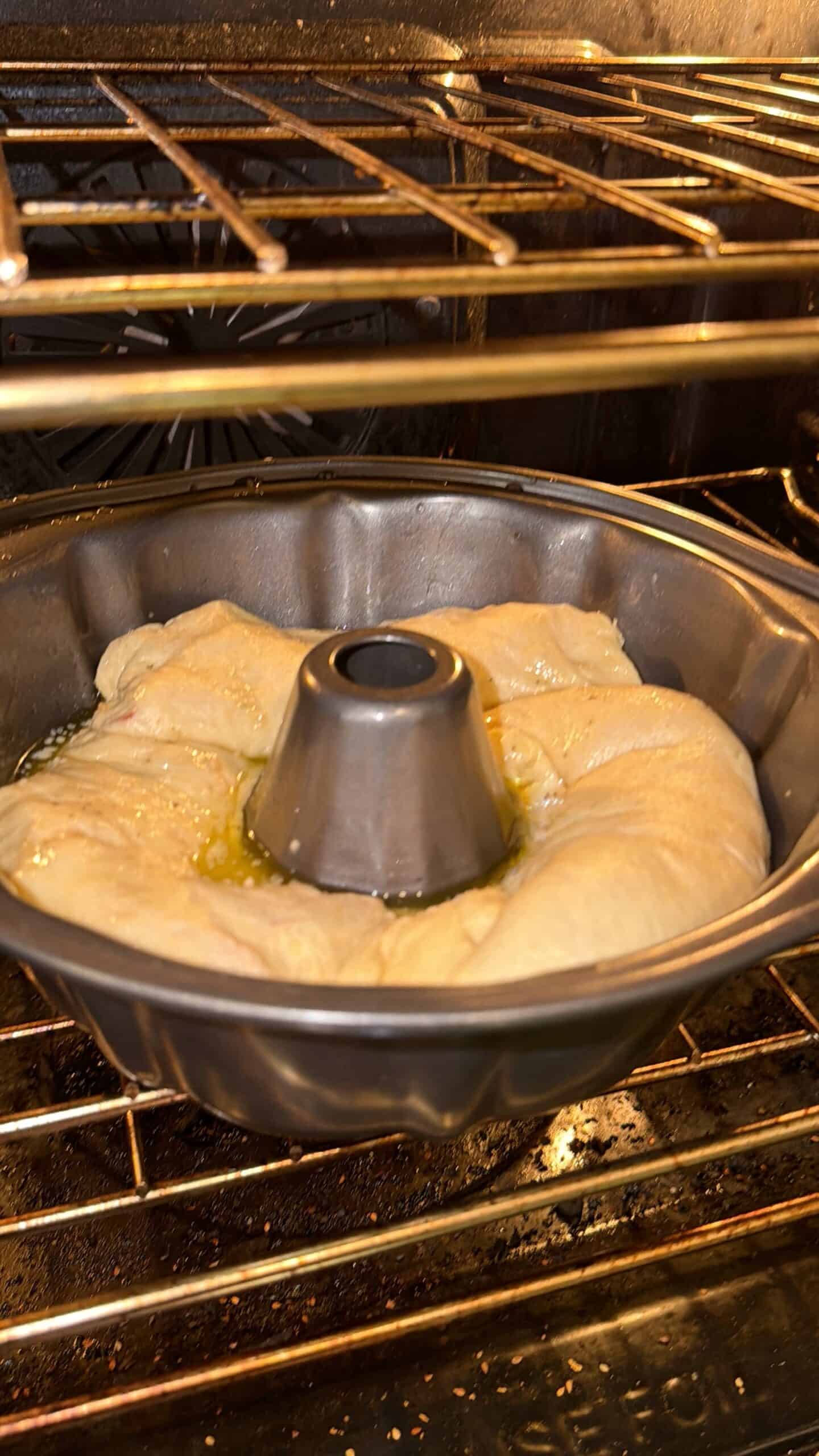 A bundt pan with dough rising inside an oven, with melted butter or oil visible on the dough’s surface, mid-bake under the oven’s warm light.