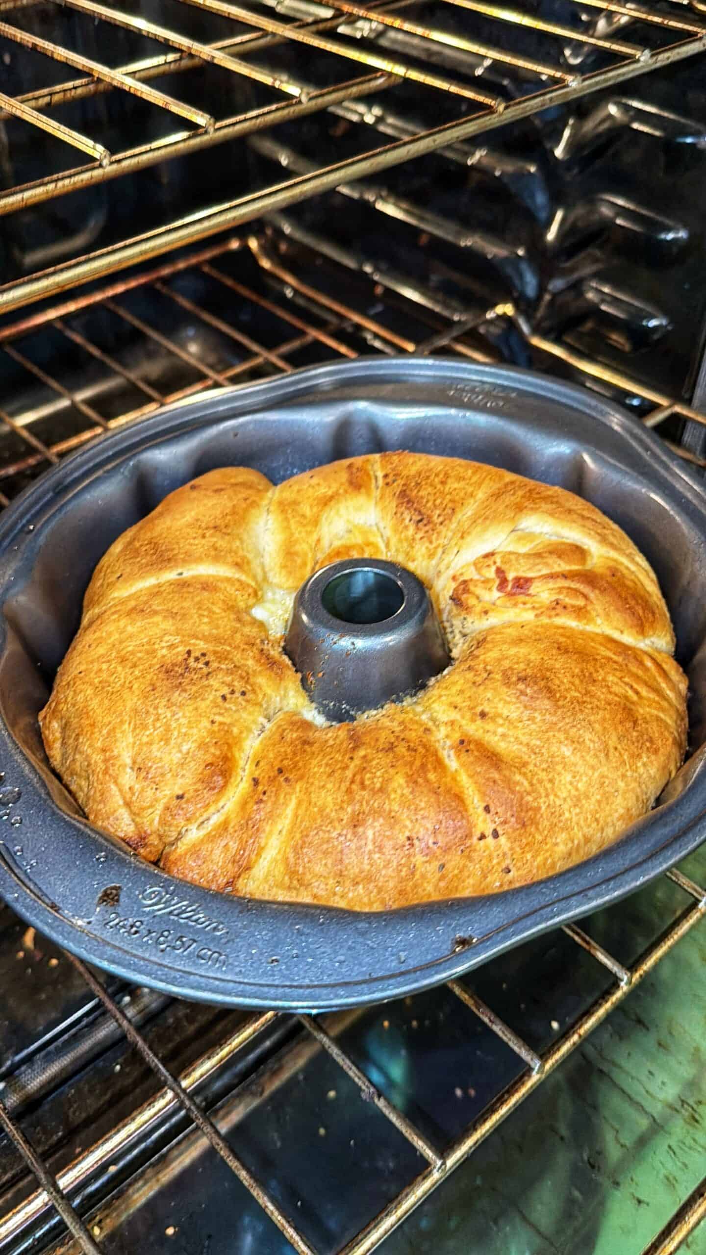A golden-brown ring of bread is baking in a Bundt pan inside an oven. The bread is puffed up and evenly browned, with a soft texture visible on its surface.