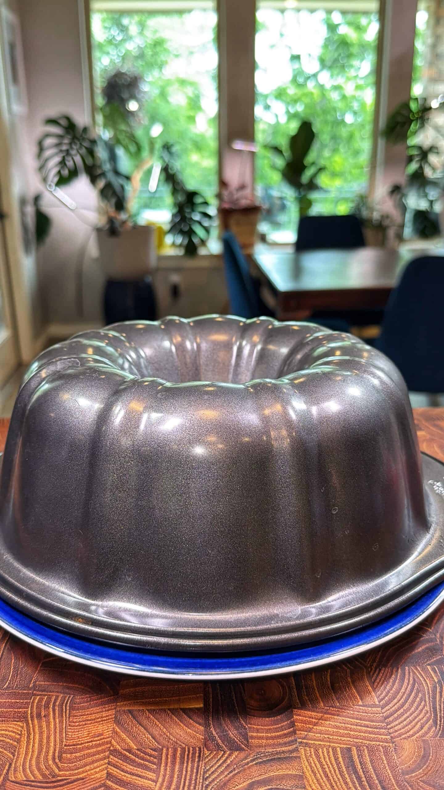 A metallic bundt cake pan sits upside down on stacked blue plates atop a wooden countertop, with a bright dining room and large green plants visible in the background.