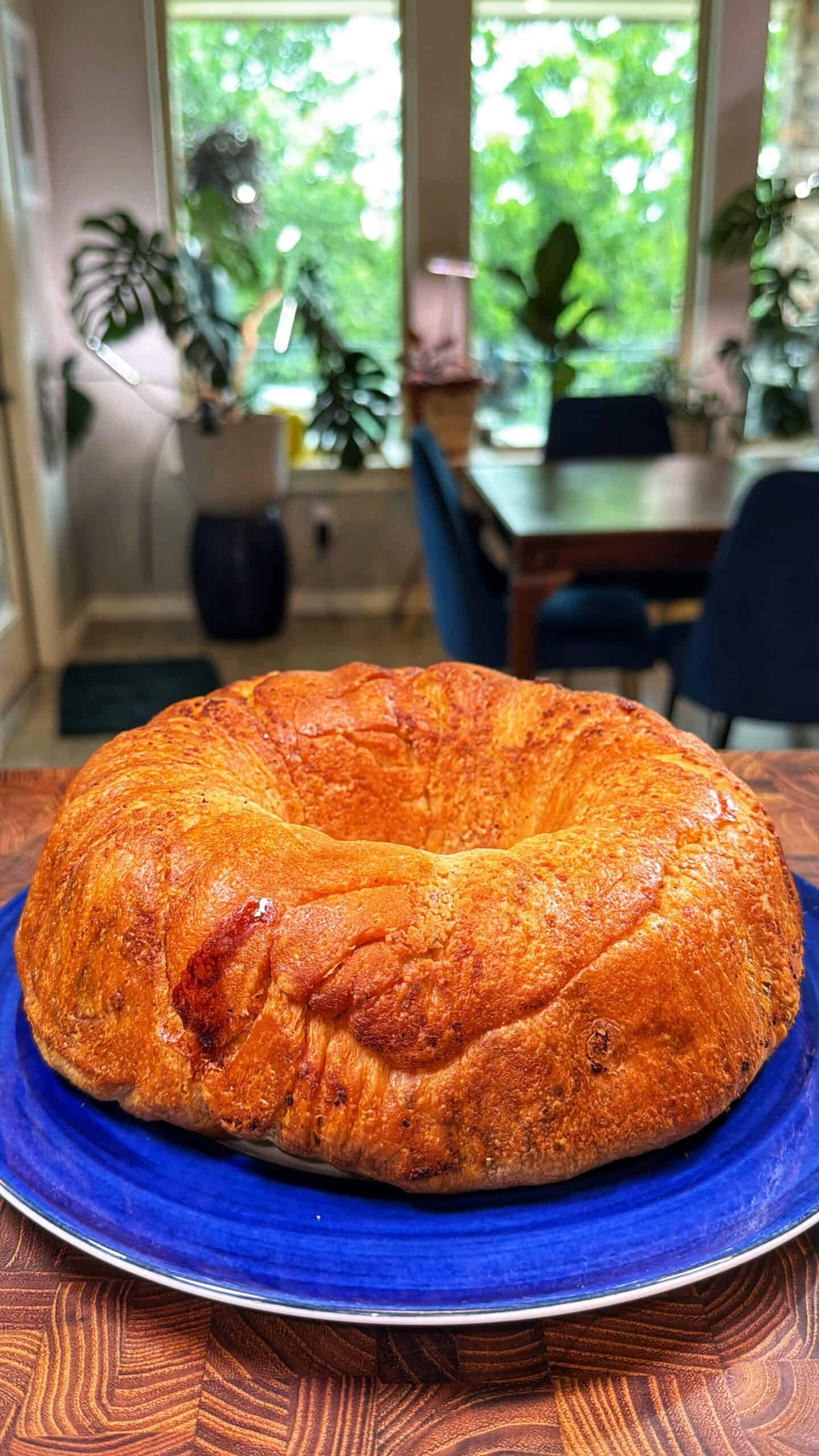 A golden-brown, ring-shaped bread sits on a blue plate. The background shows a dining table, blue chairs, and large windows with lush green plants outside.