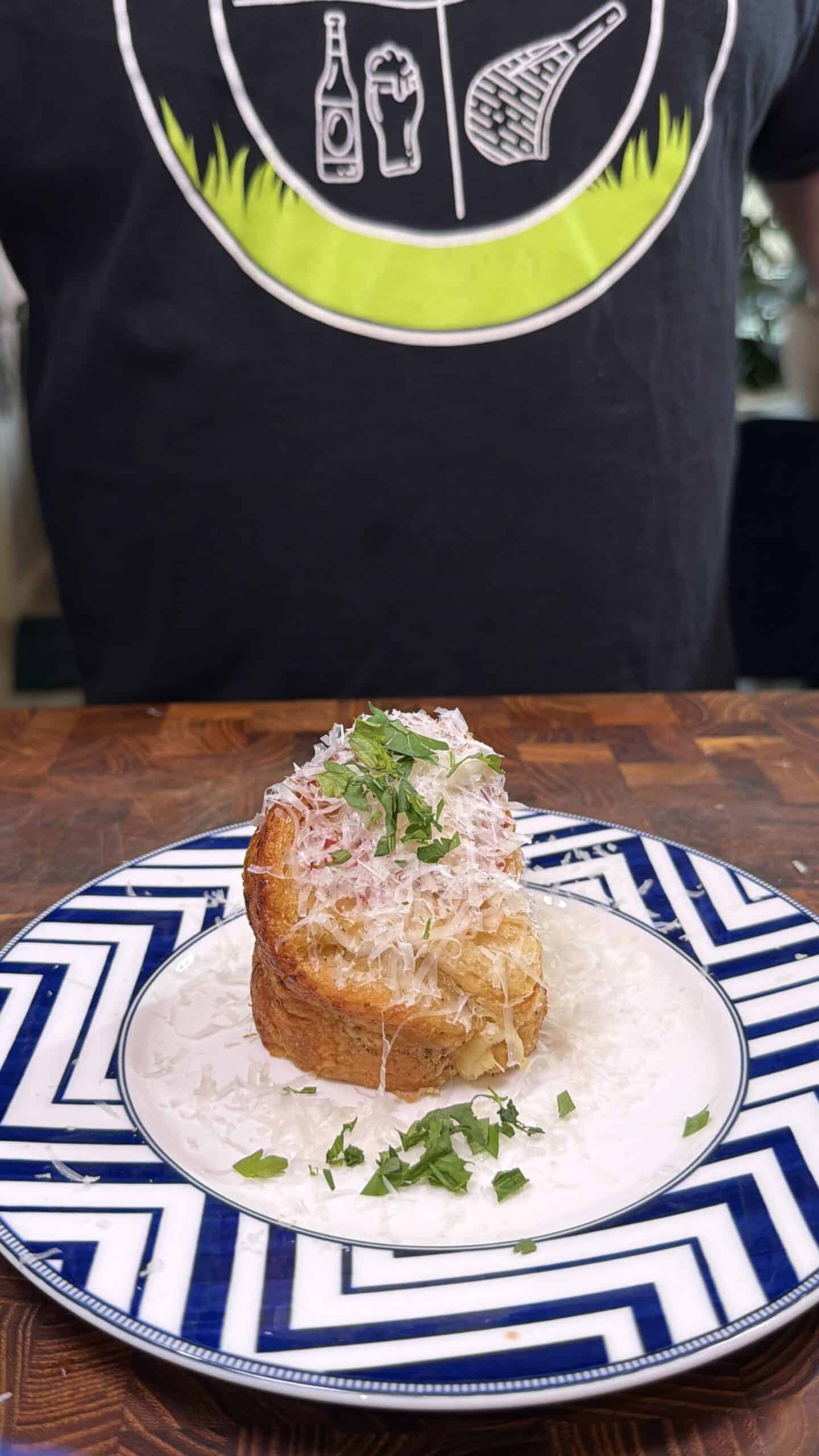 A plate with a savory muffin topped with shredded cheese and chopped parsley sits on a blue and white patterned plate. A person in a dark shirt with food illustrations stands behind the dish.