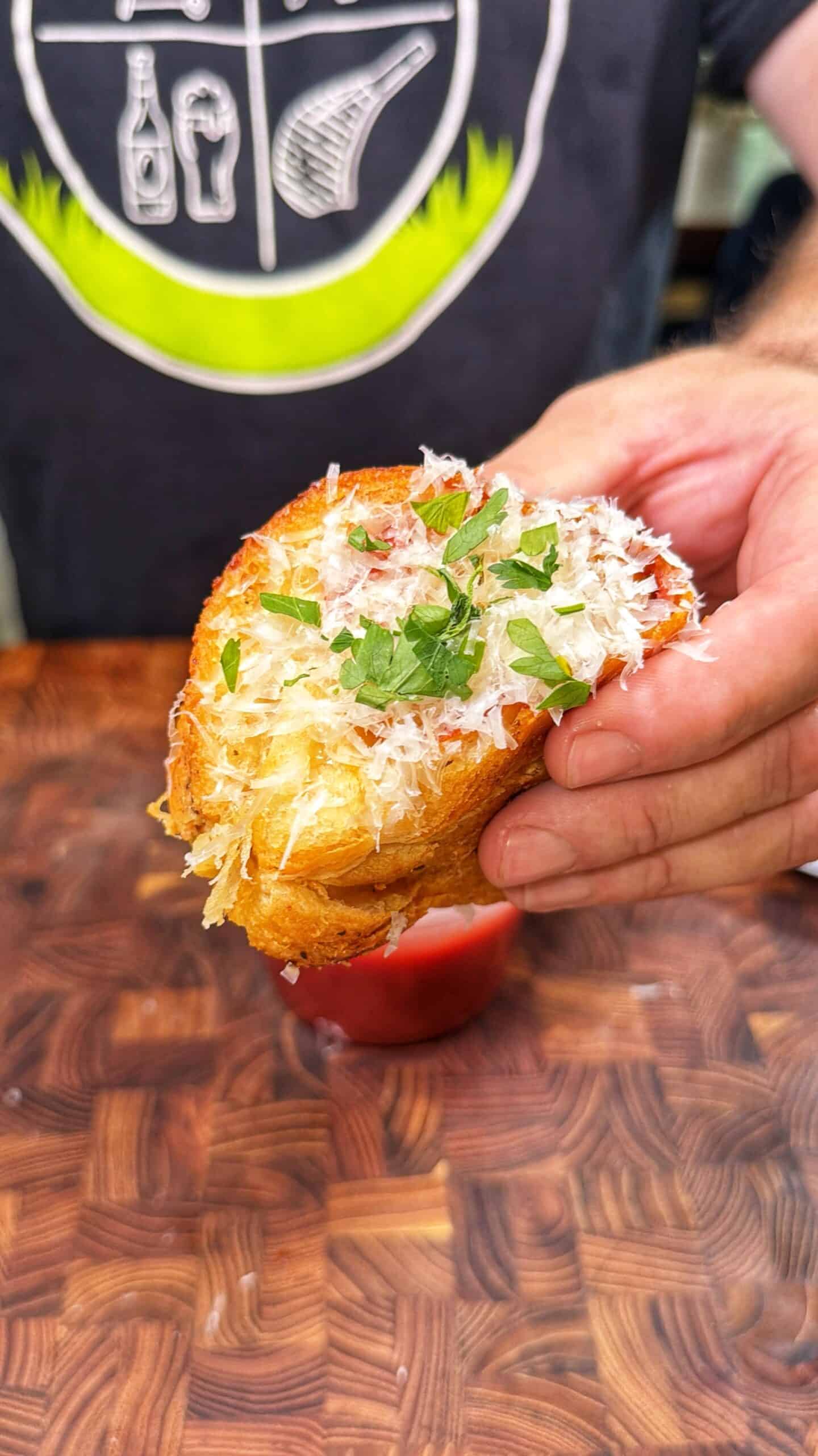 A person holds a toasted, golden bread slice topped with shredded cheese and chopped herbs, poised above a small container of red dipping sauce on a wooden surface.