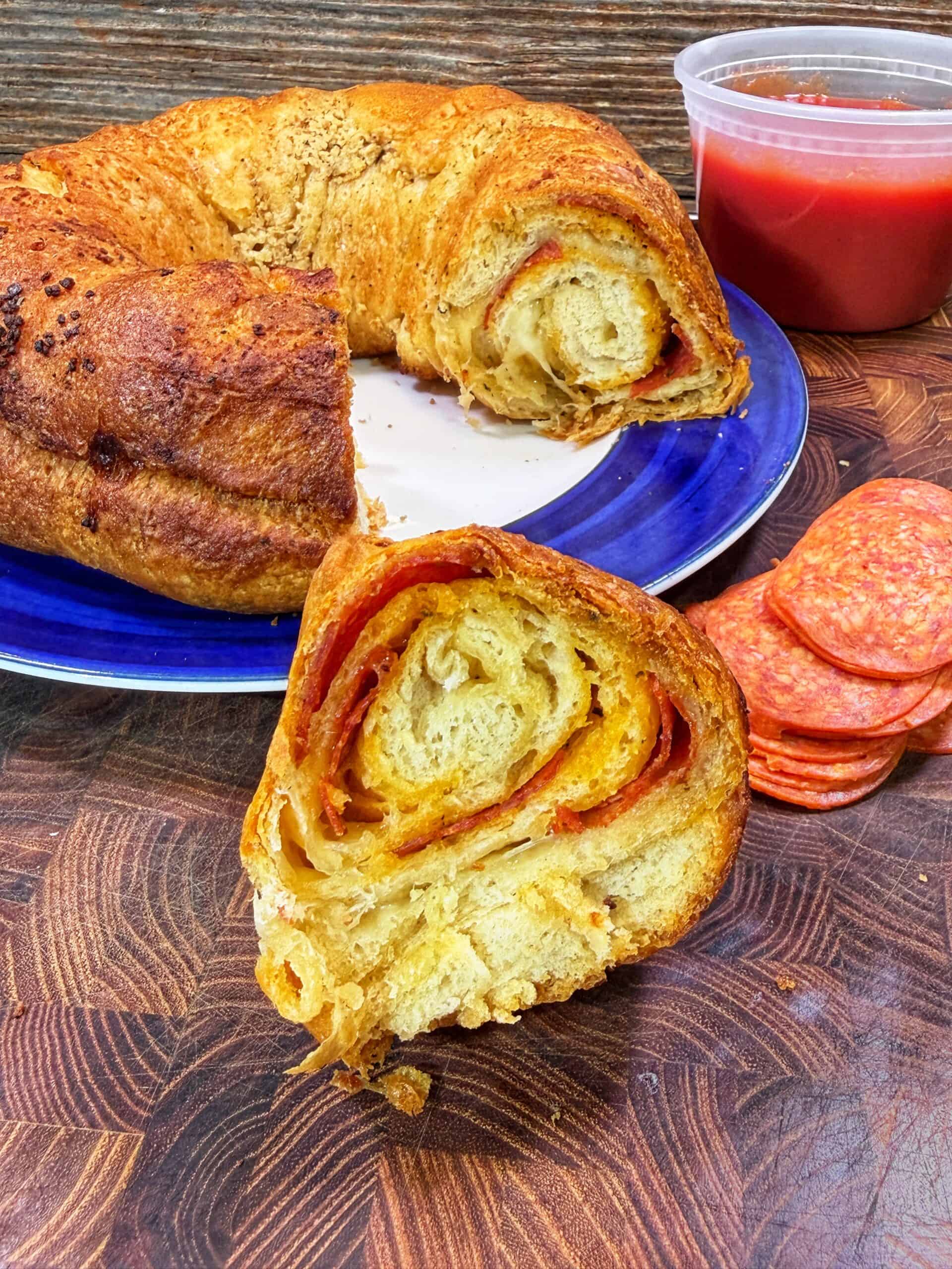 A slice of stuffed bread with layers of pepperoni and cheese sits on a wooden board, with more bread on a blue plate, a stack of pepperoni slices, and a container of red dipping sauce beside it.