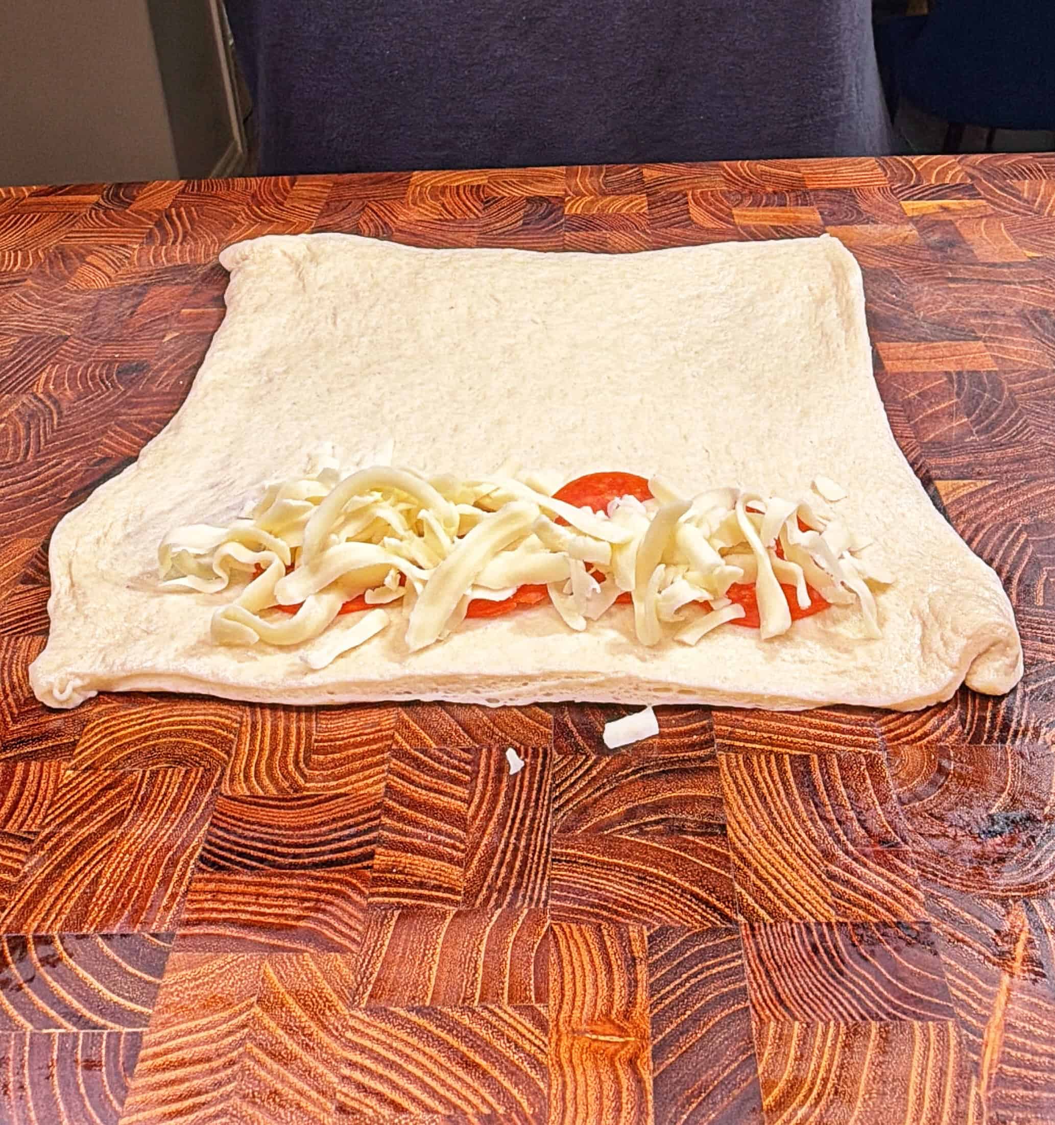 A square sheet of dough on a wooden surface with shredded cheese and tomato slices lined near one edge, ready to be rolled or folded.