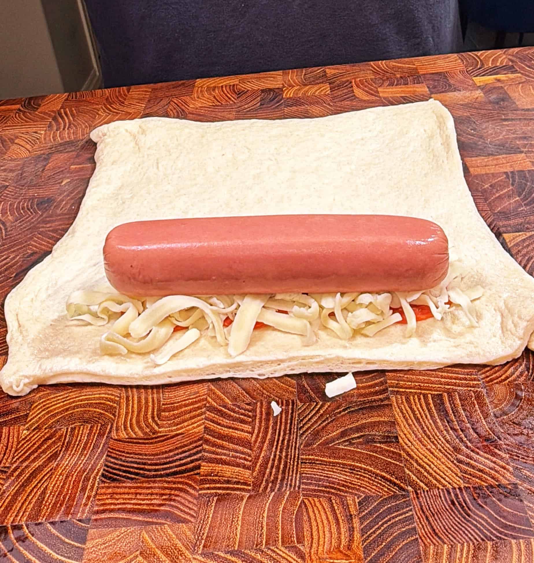 A raw hot dog placed on a layer of shredded cheese and chopped vegetables, all resting on a square piece of uncooked dough on a wooden surface.
