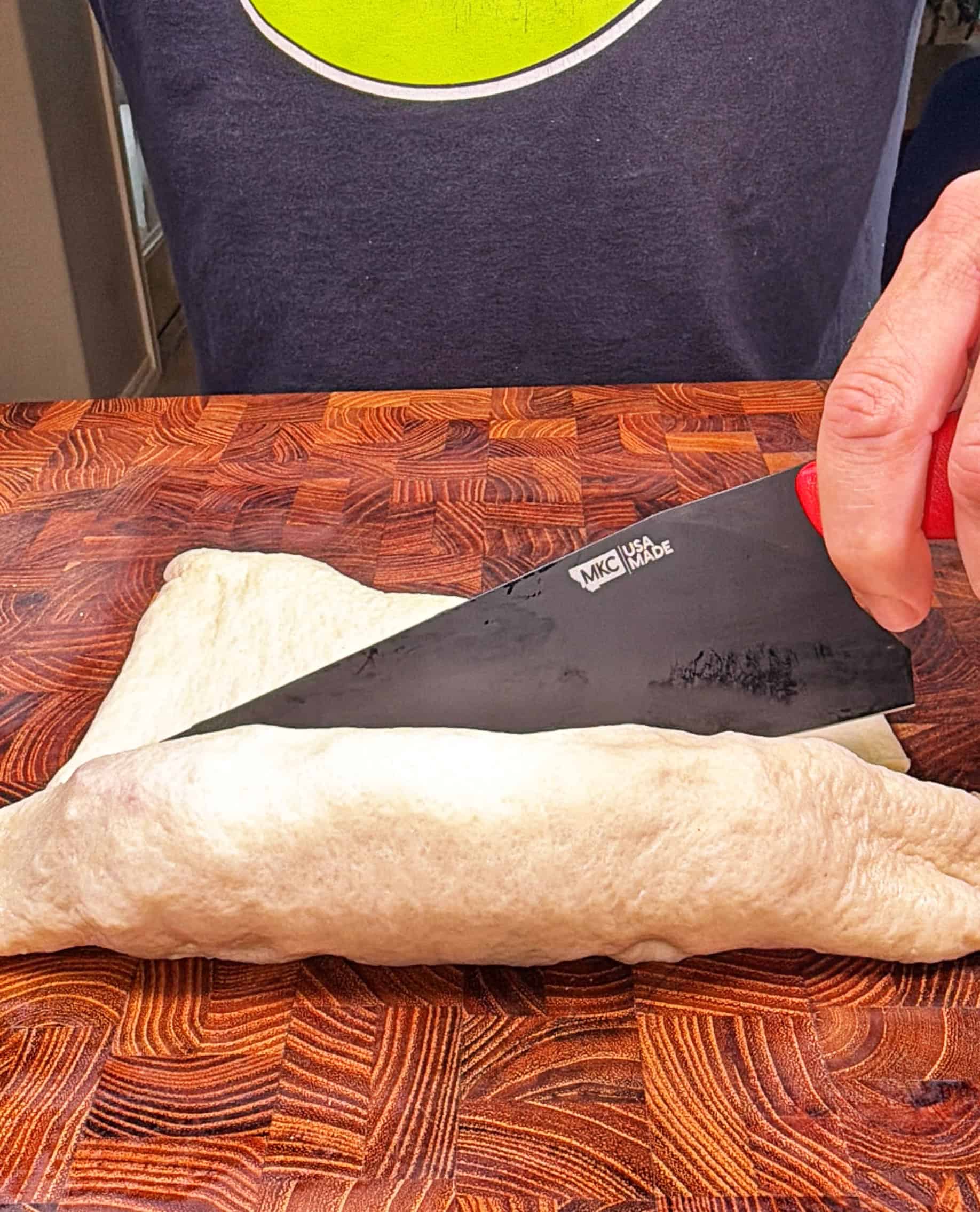 A person uses a large knife to cut a rolled piece of dough on a wooden cutting board.
