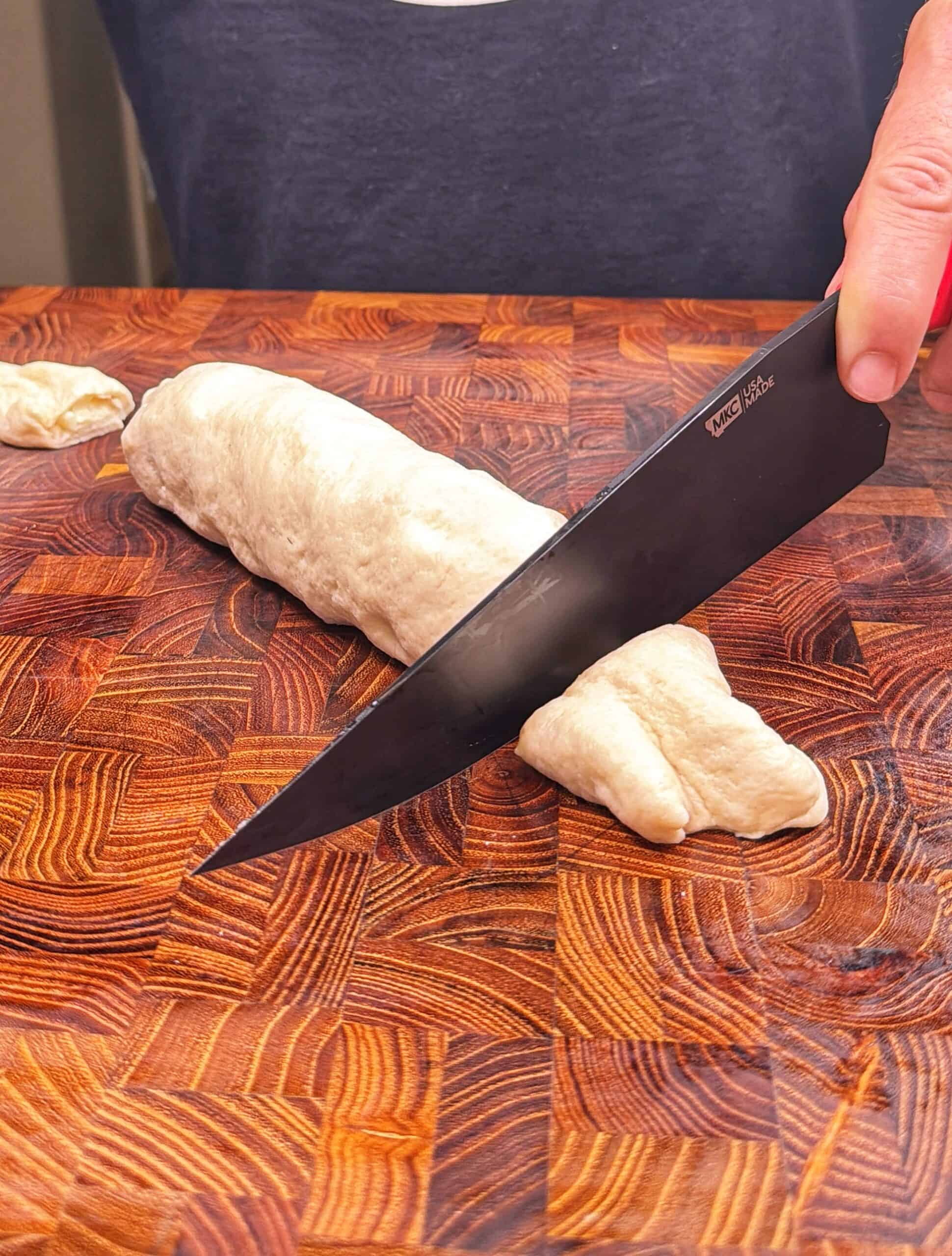 A person uses a large black knife to cut a log of dough on a wooden cutting board. The dough is partially sliced, and the surface features a detailed wood grain pattern.