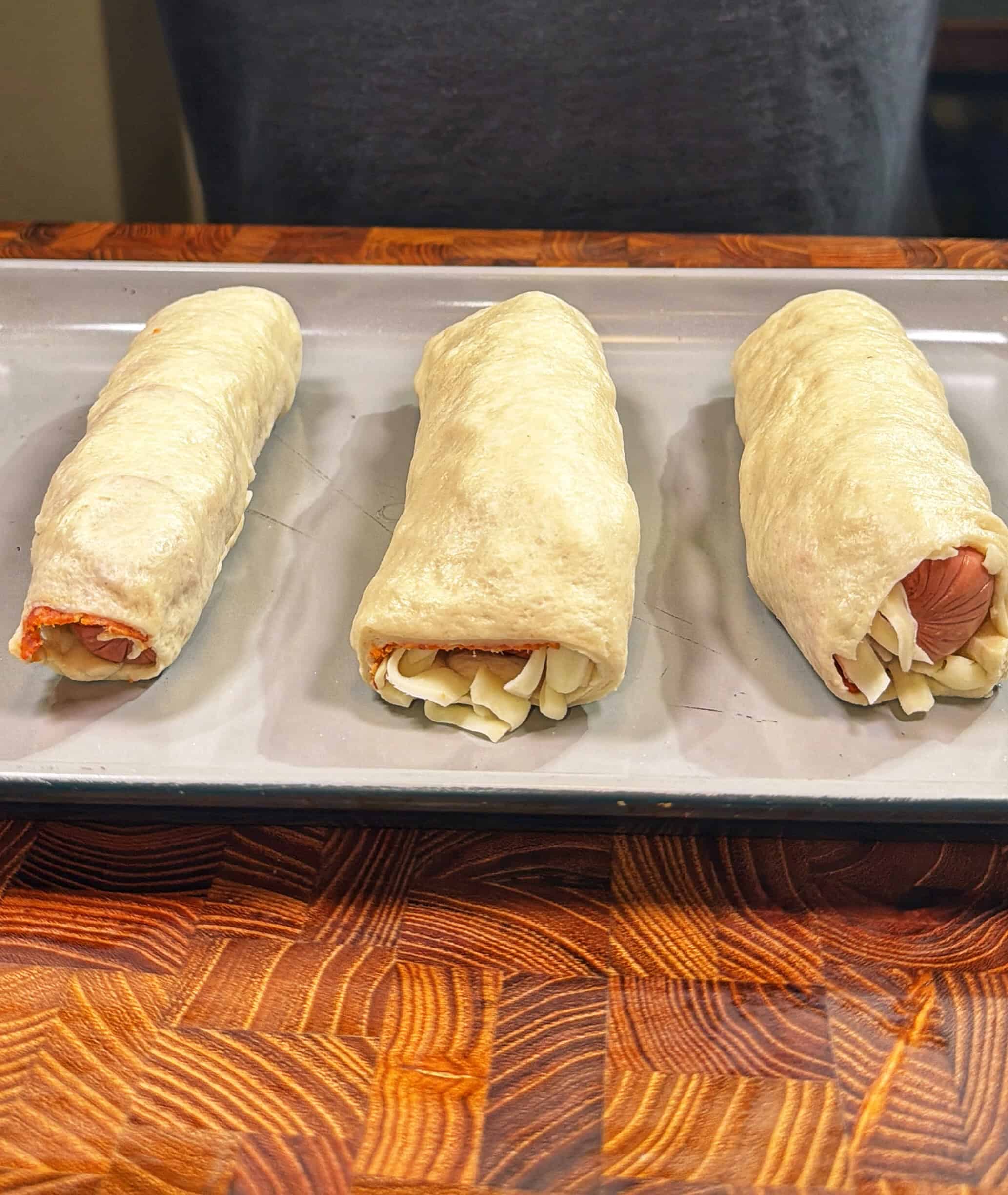 Three uncooked dough-wrapped hot dogs with cheese are placed on a baking sheet, ready to be baked. The tray sits on a wooden countertop.