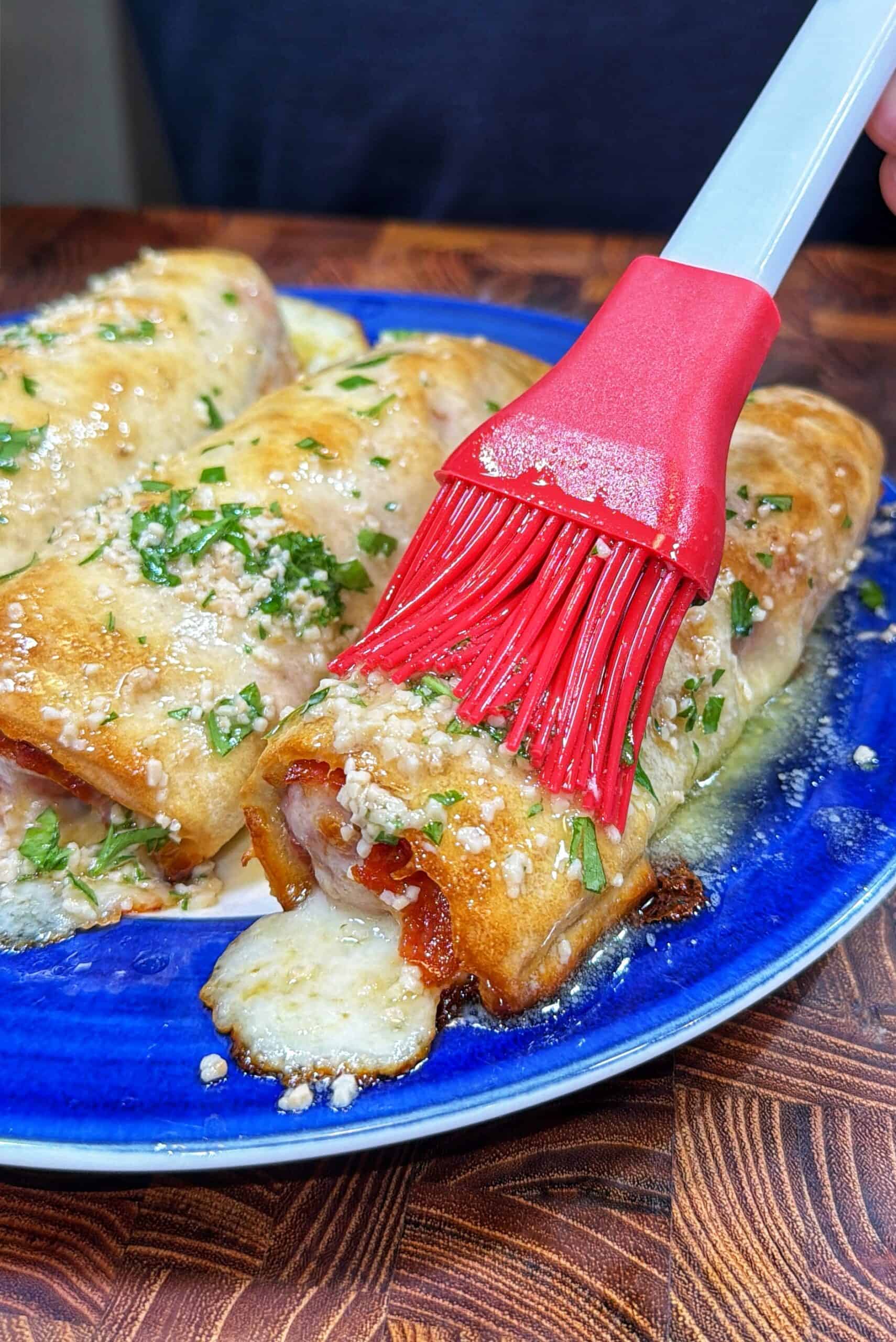 A close-up of a red brush spreading melted butter or sauce on baked, cheese-filled rolls garnished with chopped herbs and grated cheese, served on a blue plate.