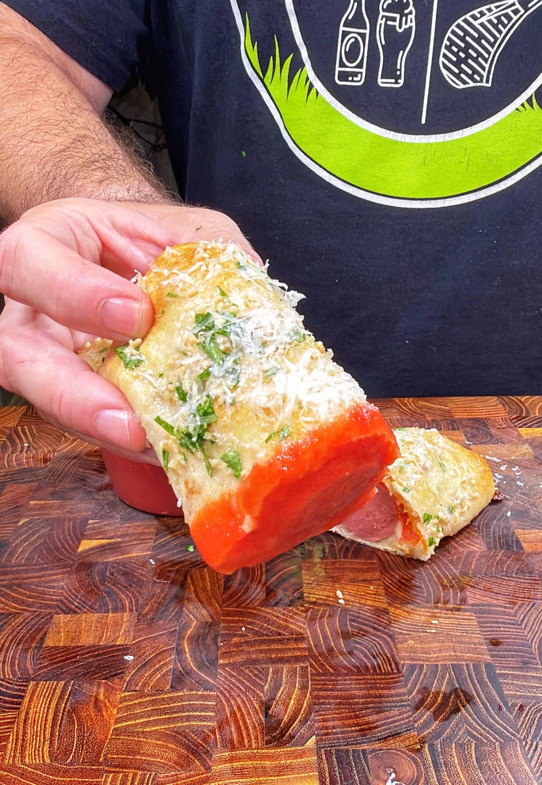 A person holds a piece of bread topped with shredded cheese, herbs, and tomato sauce. Another similar piece sits on a wooden cutting board. The person is wearing a dark shirt with a green and white graphic.