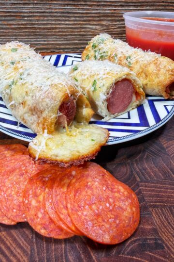 A plate holds two cheesy, baked bread rolls stuffed with hot dogs, sprinkled with grated cheese and herbs. In front are slices of pepperoni, and a container of marinara sauce is in the background.