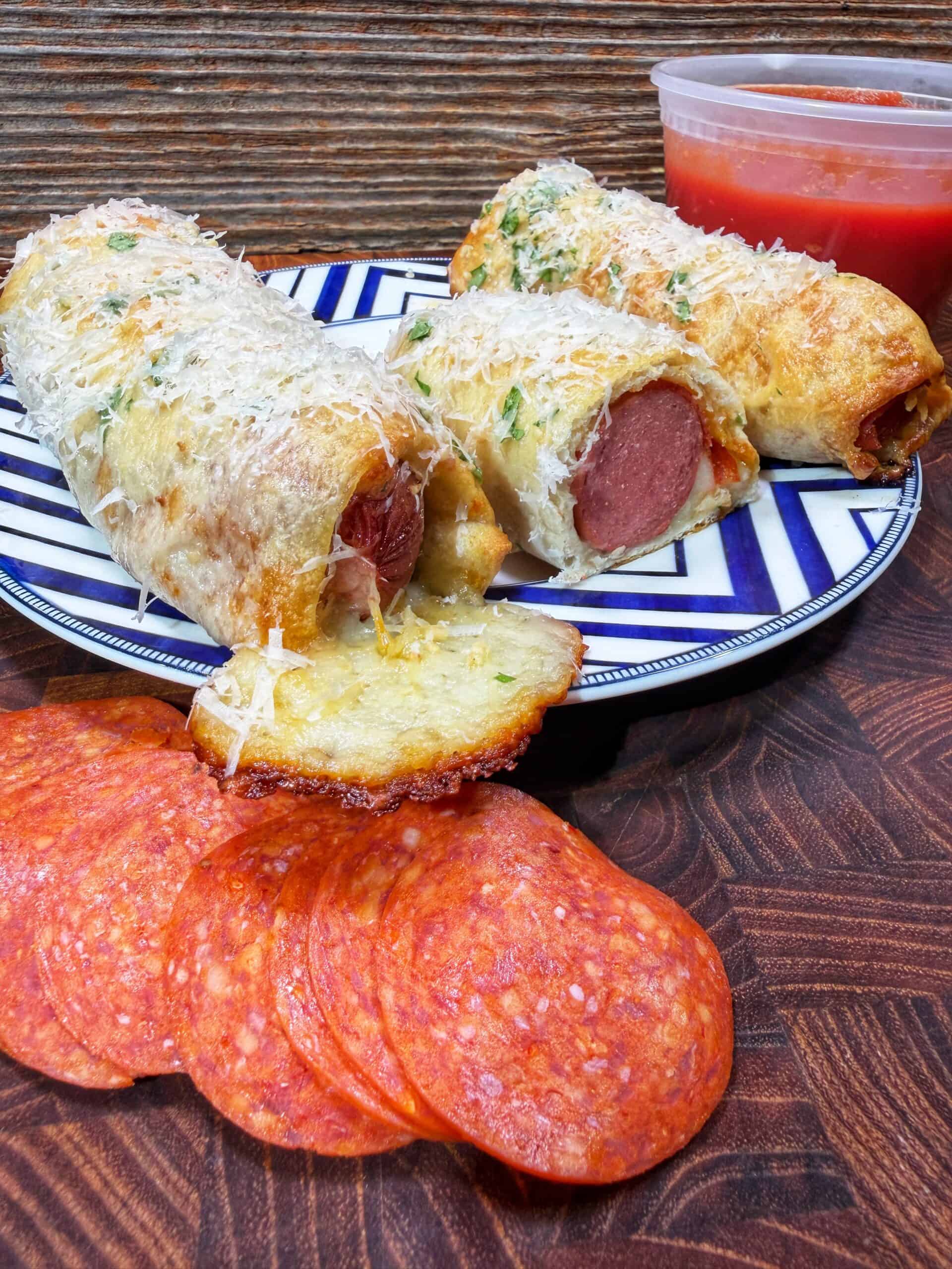 A plate holds two cheesy, baked bread rolls stuffed with hot dogs, sprinkled with grated cheese and herbs. In front are slices of pepperoni, and a container of marinara sauce is in the background.