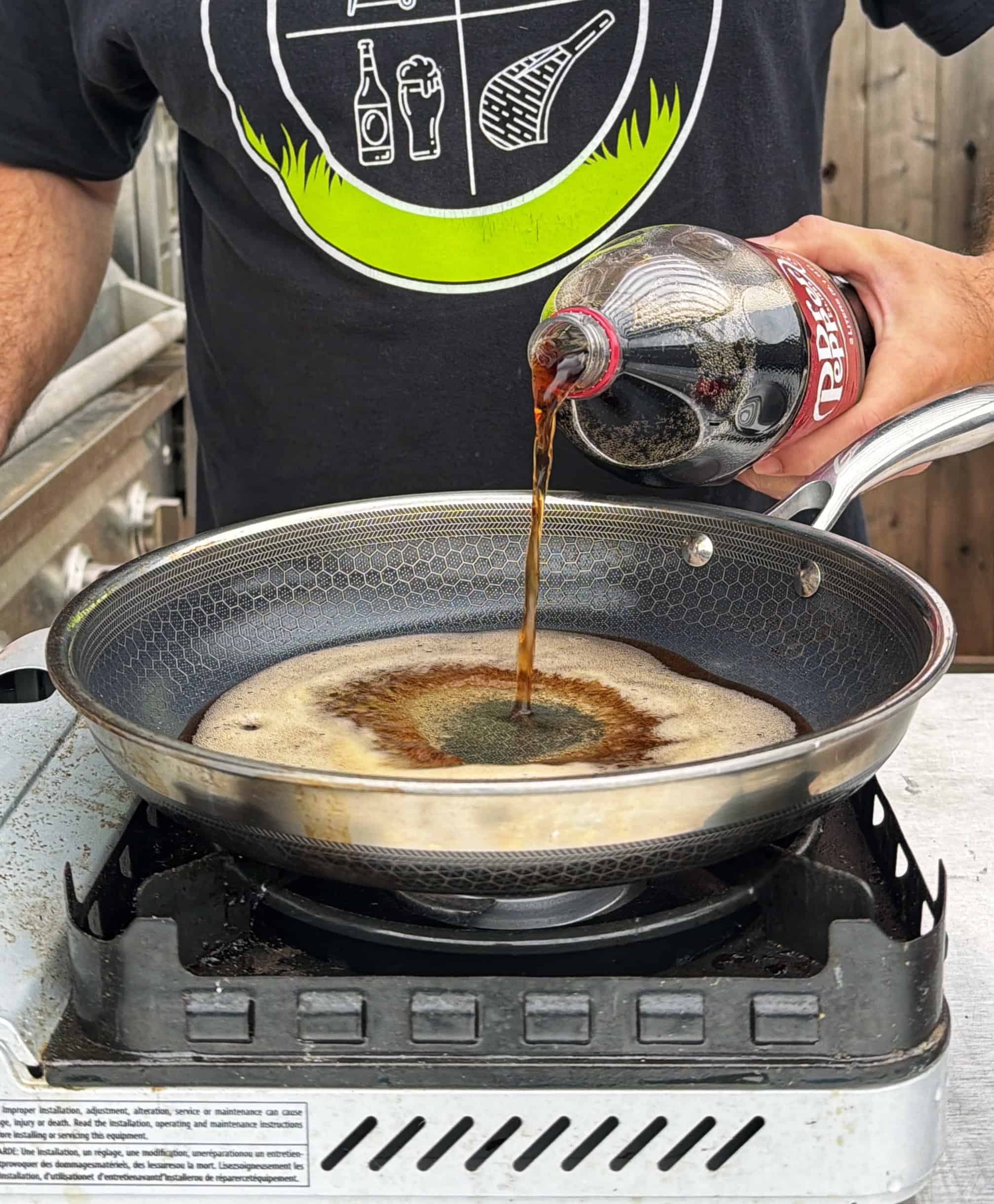 A person pours Dr Pepper soda from a bottle into a frying pan on a gas stove, wearing a black t-shirt with white and green graphics.