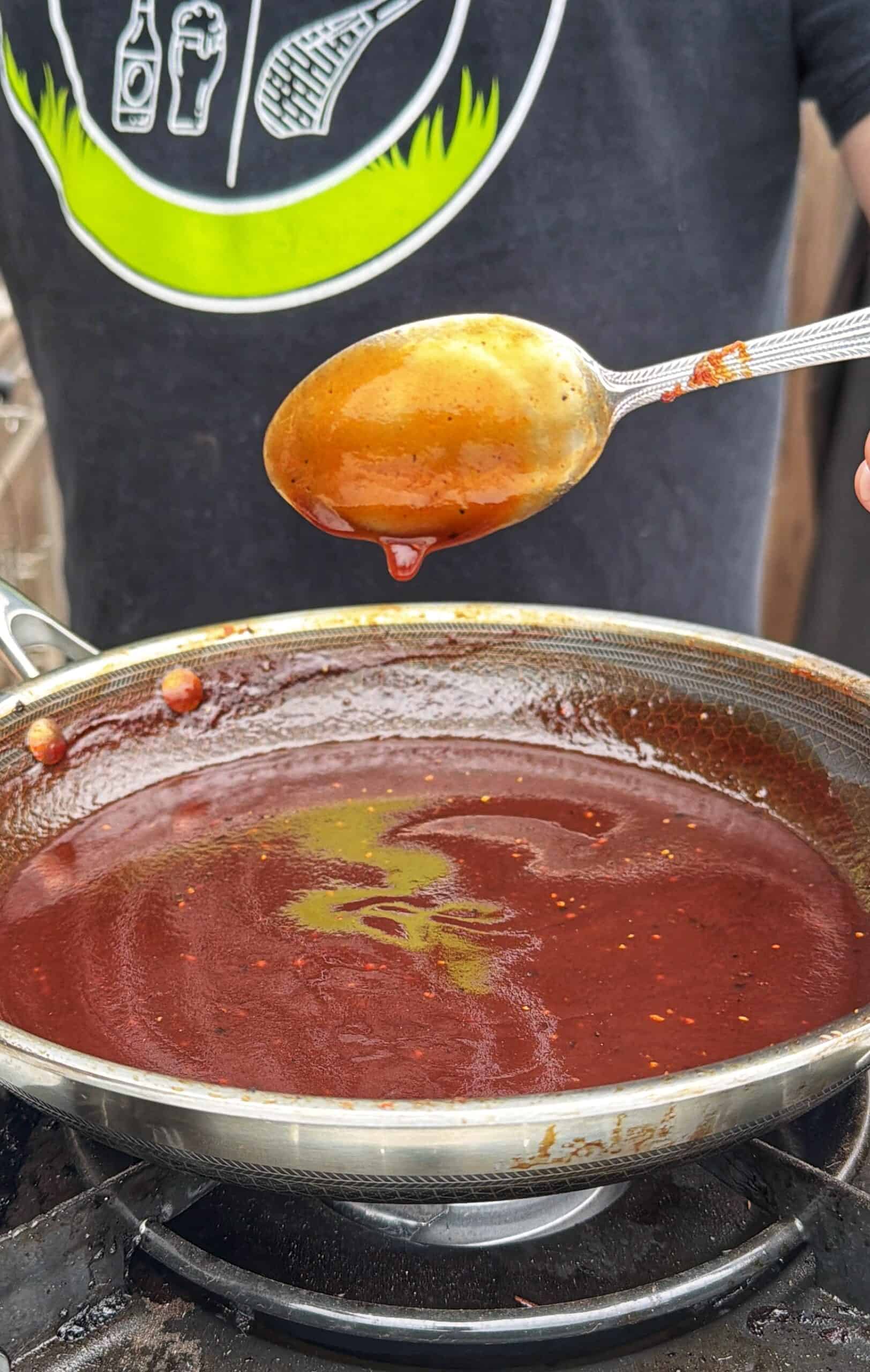 A person holds a spoon over a pan filled with dark red sauce, possibly barbecue sauce, on a stovetop. The persons shirt has a green and white graphic with food-related images.