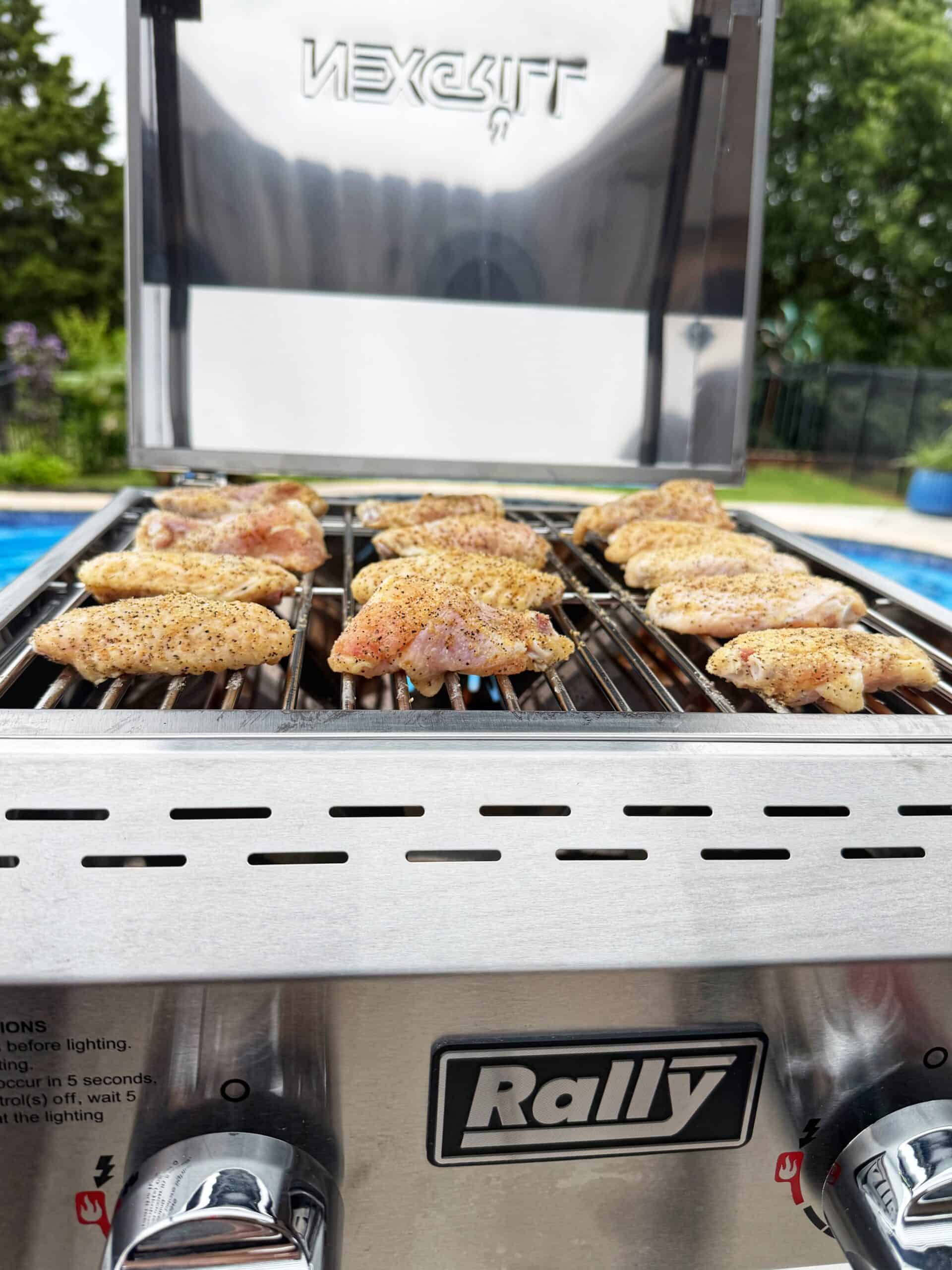 Chicken wings are cooking on a stainless steel Nexgrill gas grill with the Rally badge, set outdoors next to a swimming pool and surrounded by greenery.