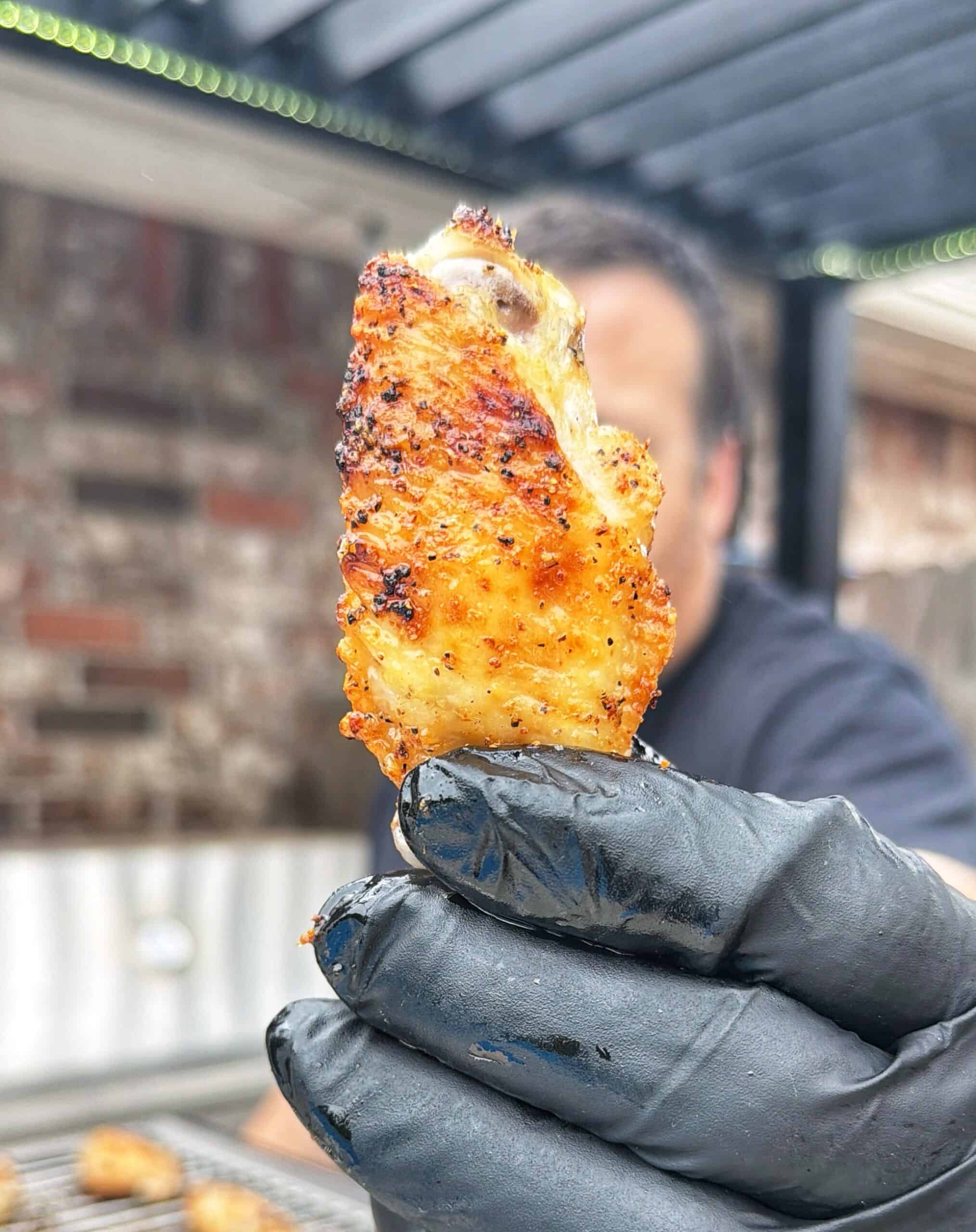 A person wearing a black glove holds up a grilled, seasoned chicken wing in focus, with an outdoor grill and blurred background visible.