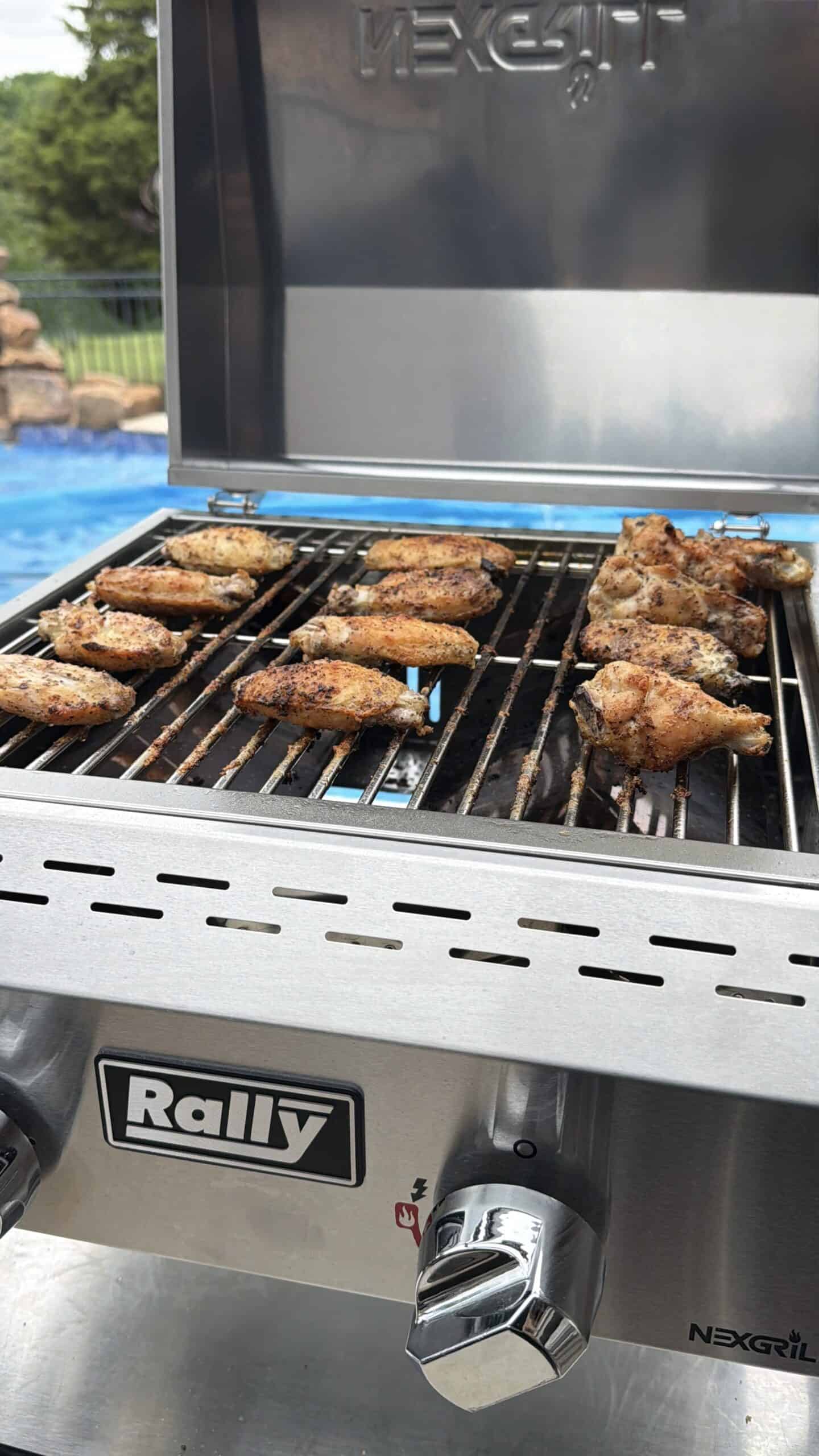 Chicken wings grilling on a stainless steel Rally Nexgrill barbecue with the lid open, next to a swimming pool and greenery in the background.