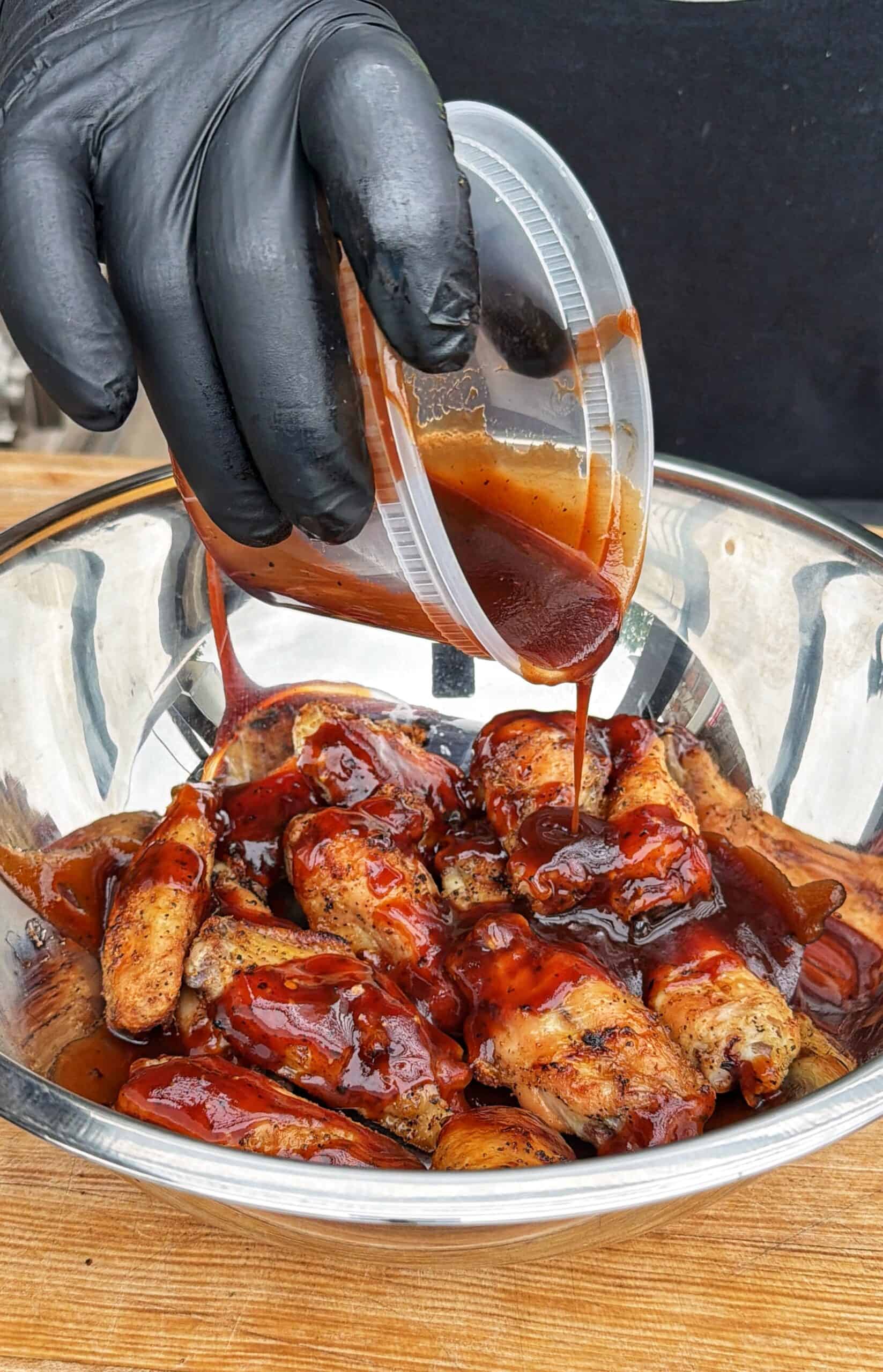 A gloved hand pours barbecue sauce from a plastic container onto grilled chicken wings in a large metal mixing bowl.