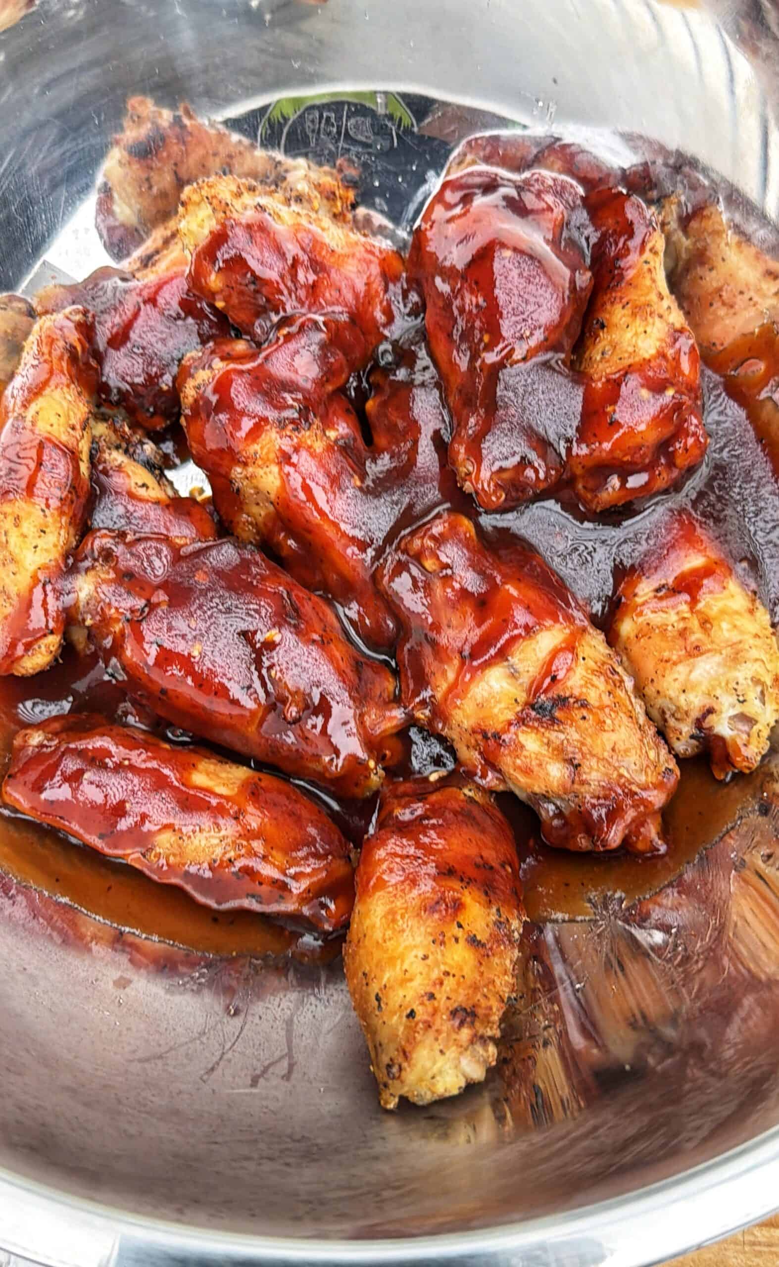 A close-up of chicken wings coated in a glossy barbecue sauce, arranged in a metal bowl. The wings are golden brown and generously covered in the thick, reddish sauce.