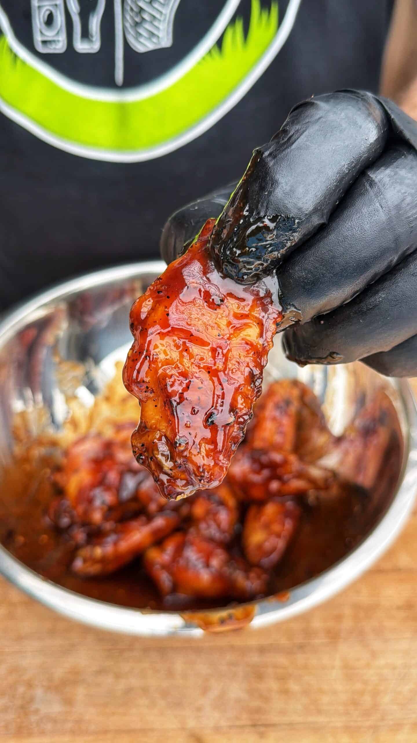 A gloved hand holds a saucy, glazed chicken wing above a metal bowl filled with more chicken wings. The person wears a black shirt with a green and white design in the background.