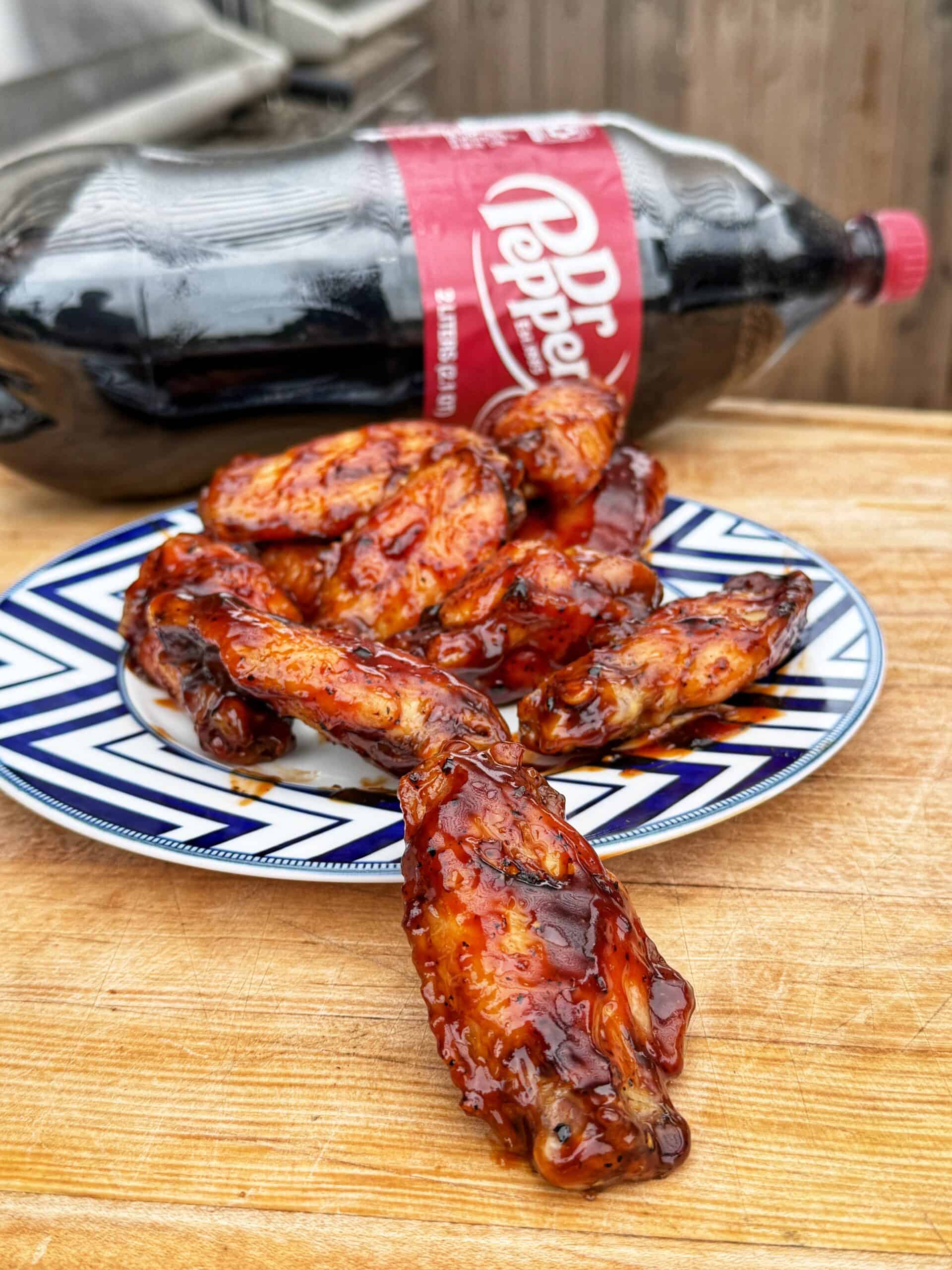 A plate of glazed chicken wings on a wooden surface, with a 2-liter bottle of Dr Pepper in the background. The wings appear sticky and well-coated in sauce.