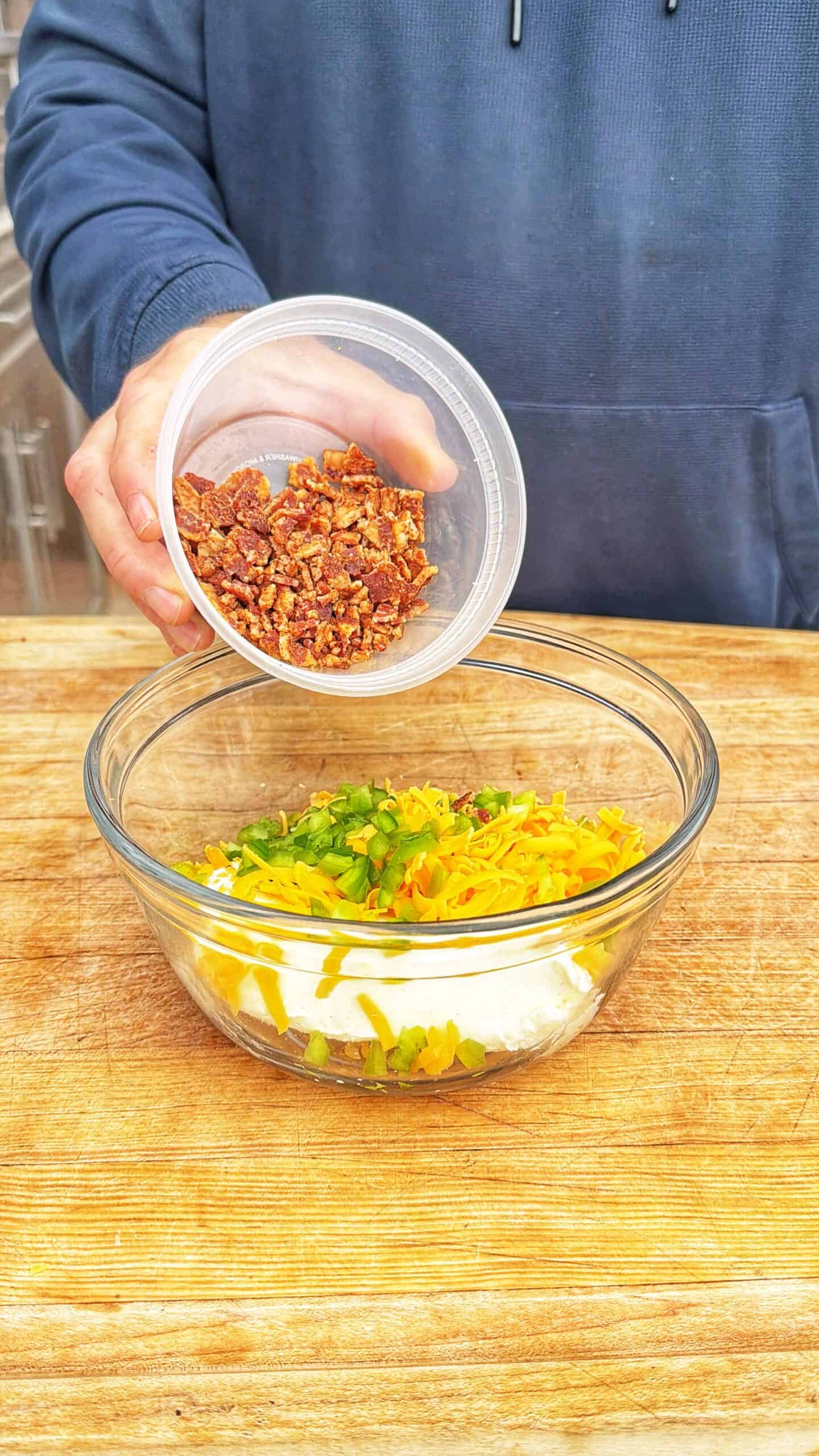 A person pours chopped bacon from a plastic container into a glass bowl containing shredded cheese, sliced green onions, and cream cheese on a wooden surface.