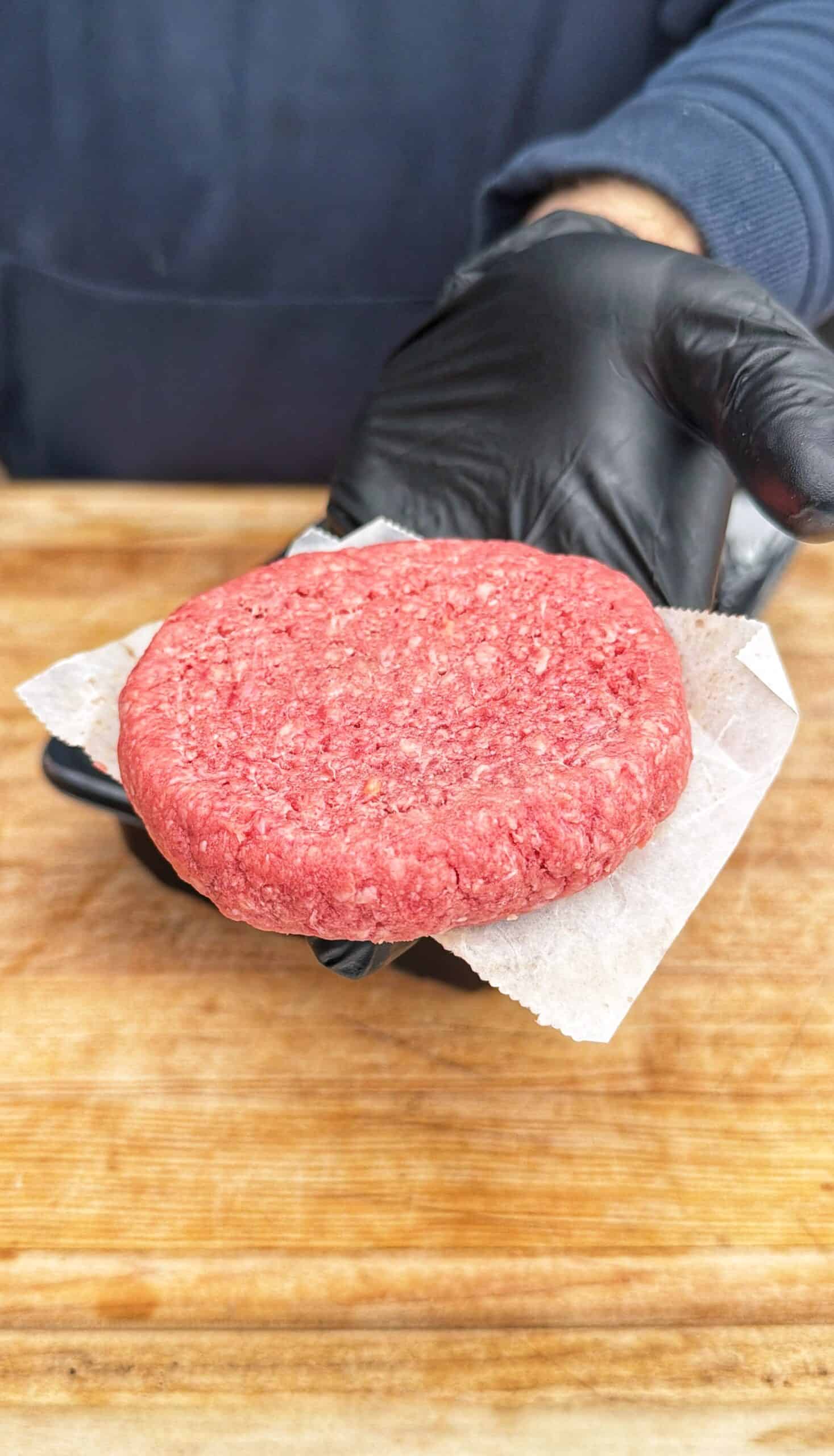 A person wearing a black glove holds a raw beef burger patty on a piece of parchment paper over a wooden surface.